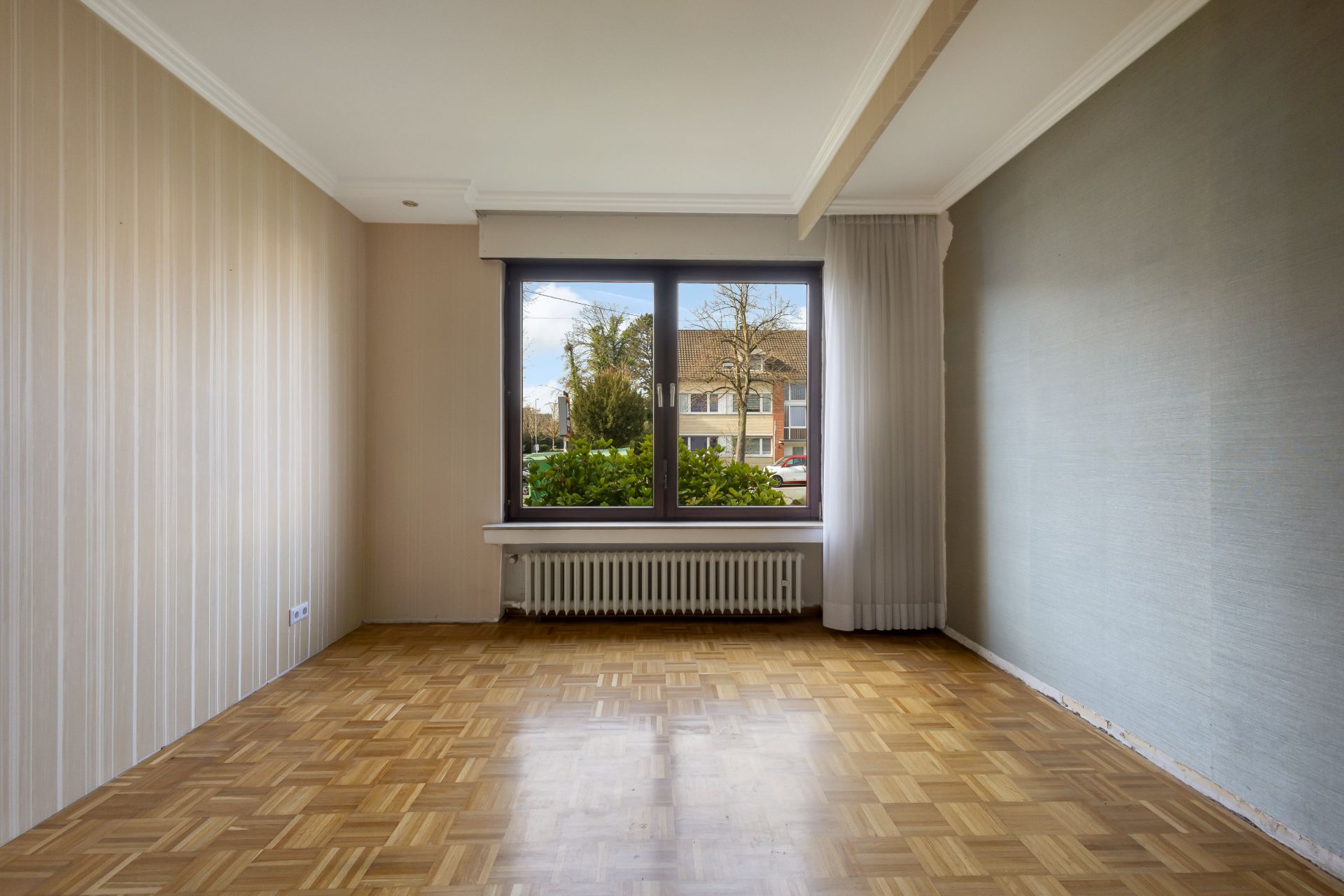Leerer Raum mit Parkettboden, großem Fenster und Heizung darunter, Blick nach draußen.