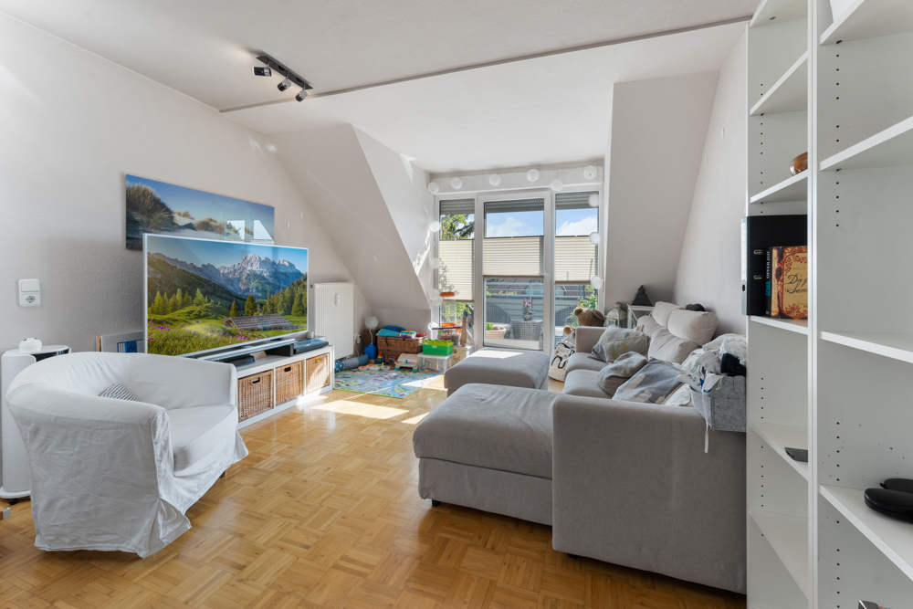 Helles Wohnzimmer mit Sofa, Sessel, Fernseher, Regal und Zugang zu einem Balkon.