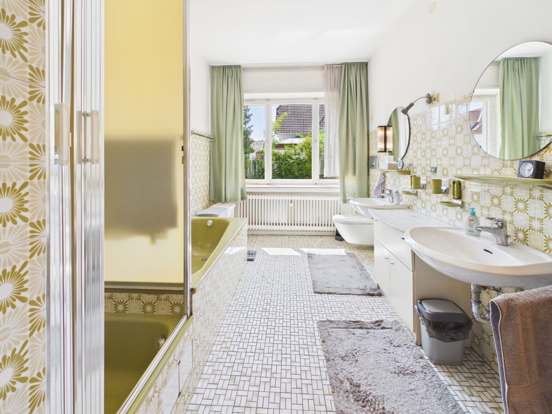 Helles Badezimmer mit Badewanne, zwei Waschbecken, Spiegel und Fenster mit grünen Vorhängen.