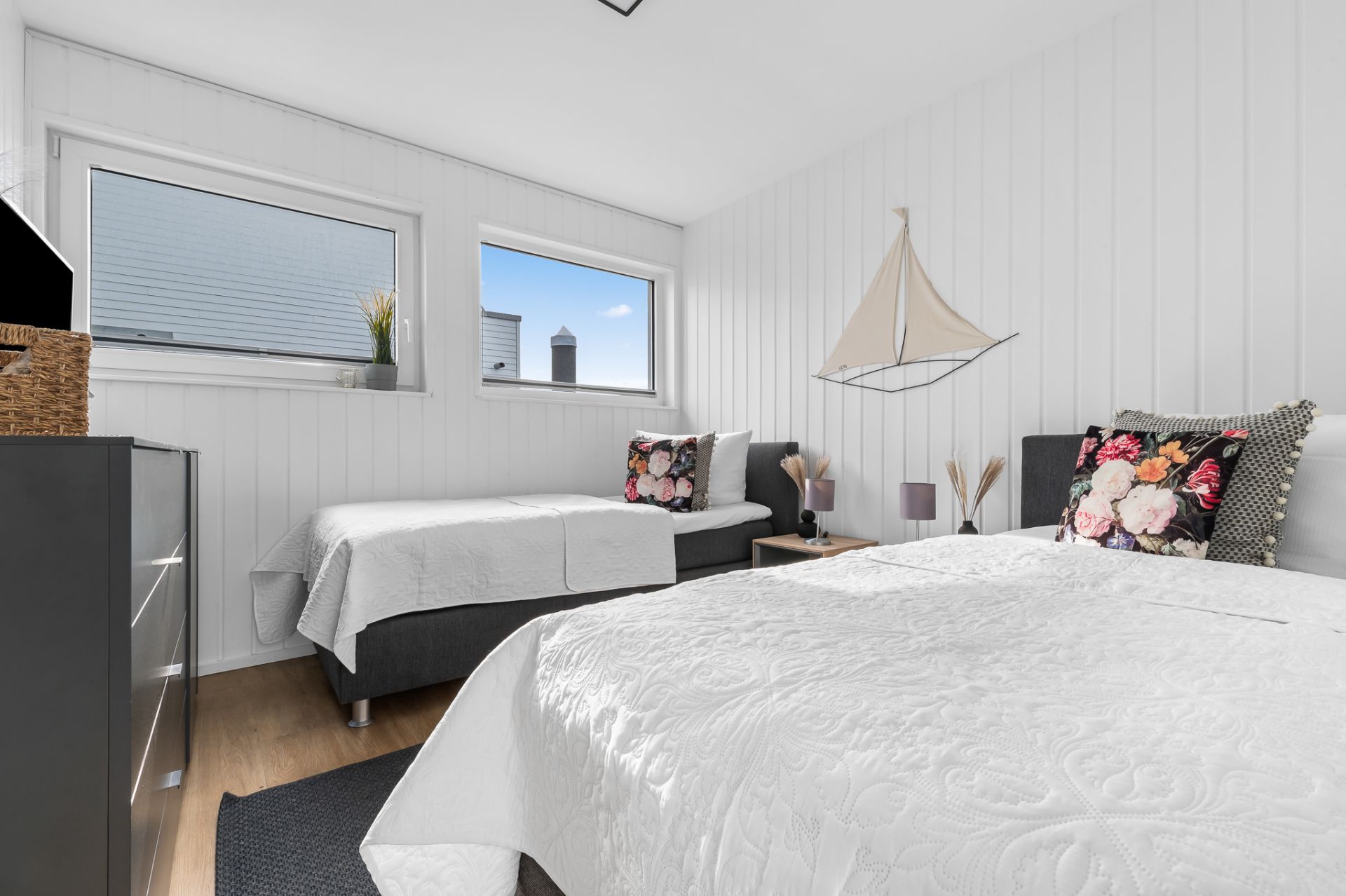 Helles Schlafzimmer mit zwei Betten, weißen Wänden und bunten Kissen, dekoriert mit einem Segelboot an der Wand.