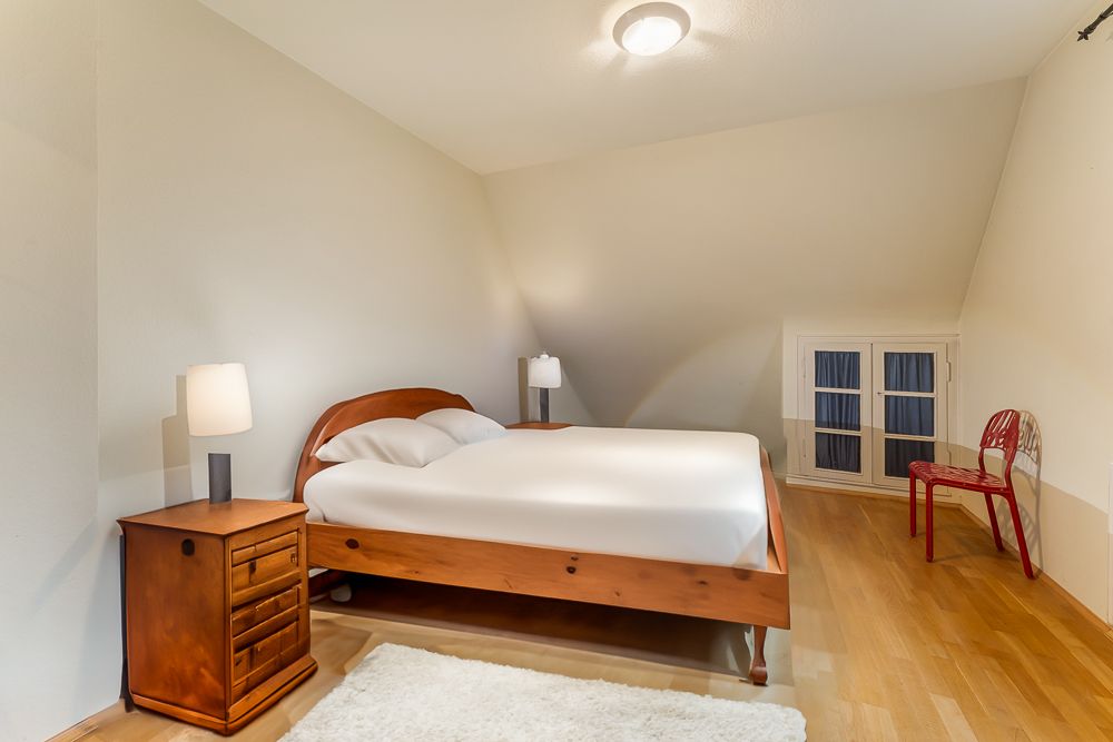Schlafzimmer mit Holzbett, Nachttisch, zwei Lampen und rotem Stuhl auf hellem Holzboden.
