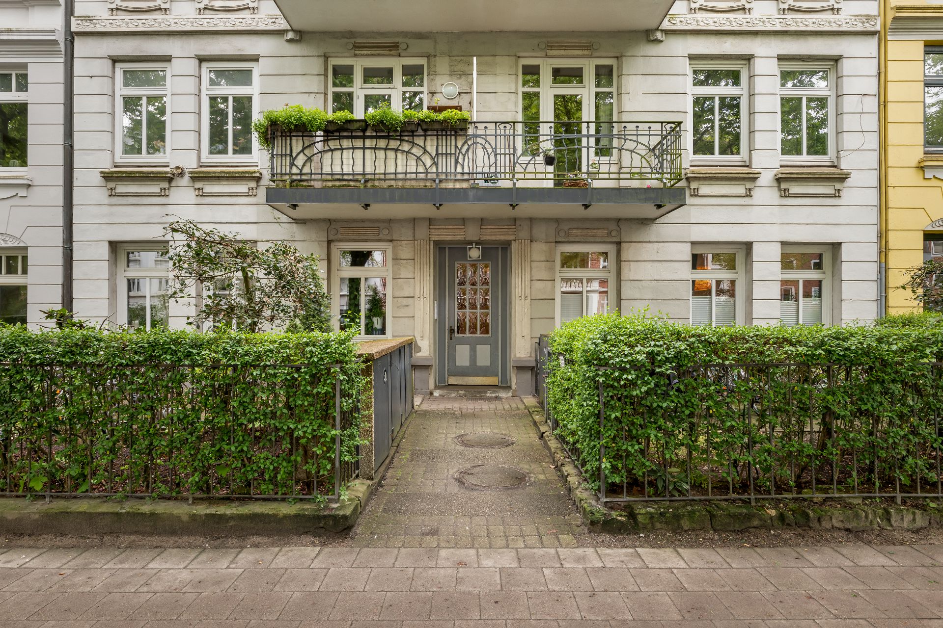 Eingang eines Wohnhauses mit Balkon, vielen Fenstern und grüner Hecke vor dem Gebäude.