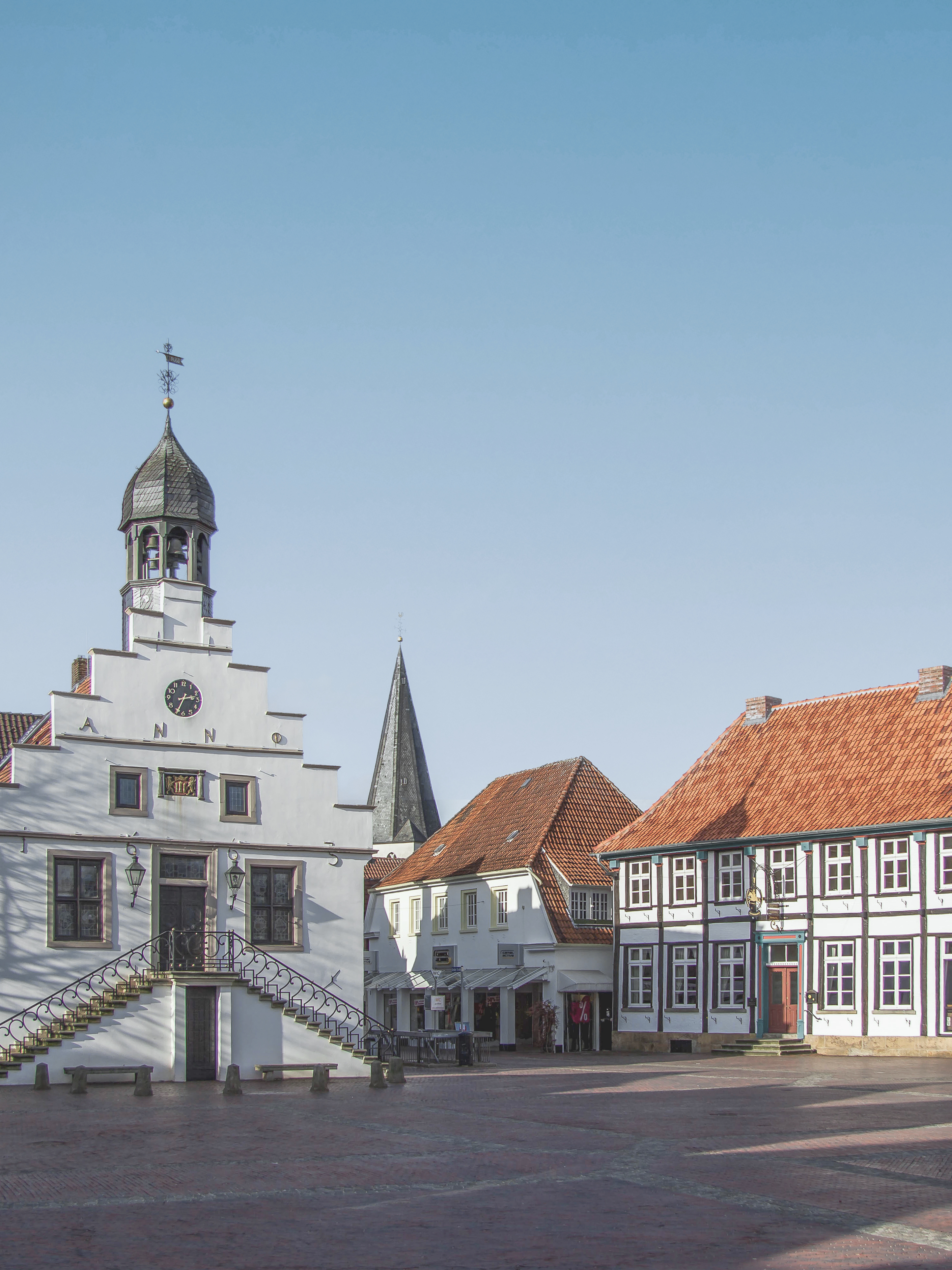Historische Gebäude mit roten Dächern und einem weißen Turm an einem sonnigen Platz.