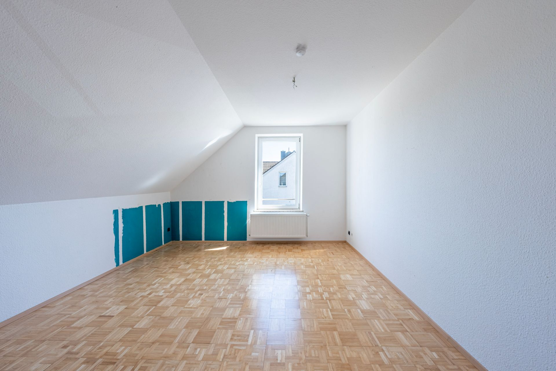 Leerer Dachgeschossraum mit Parkettboden, weiß gestrichenen Wänden und einem Fenster am Ende.