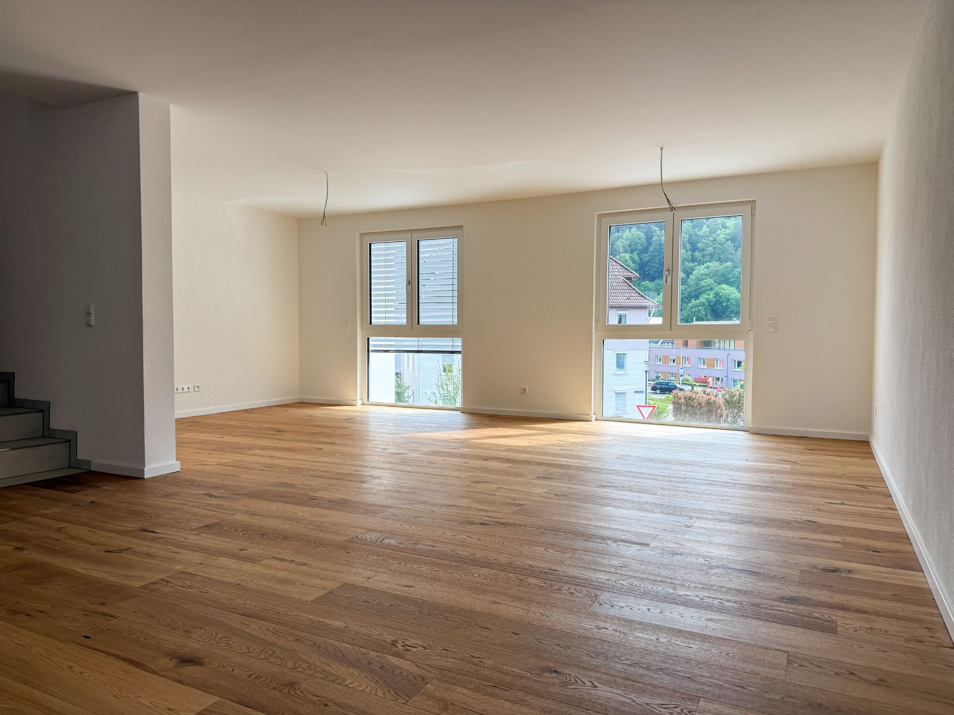 Heller, leerer Raum mit Holzfußboden, zwei großen Fenstern und einer Treppe links.