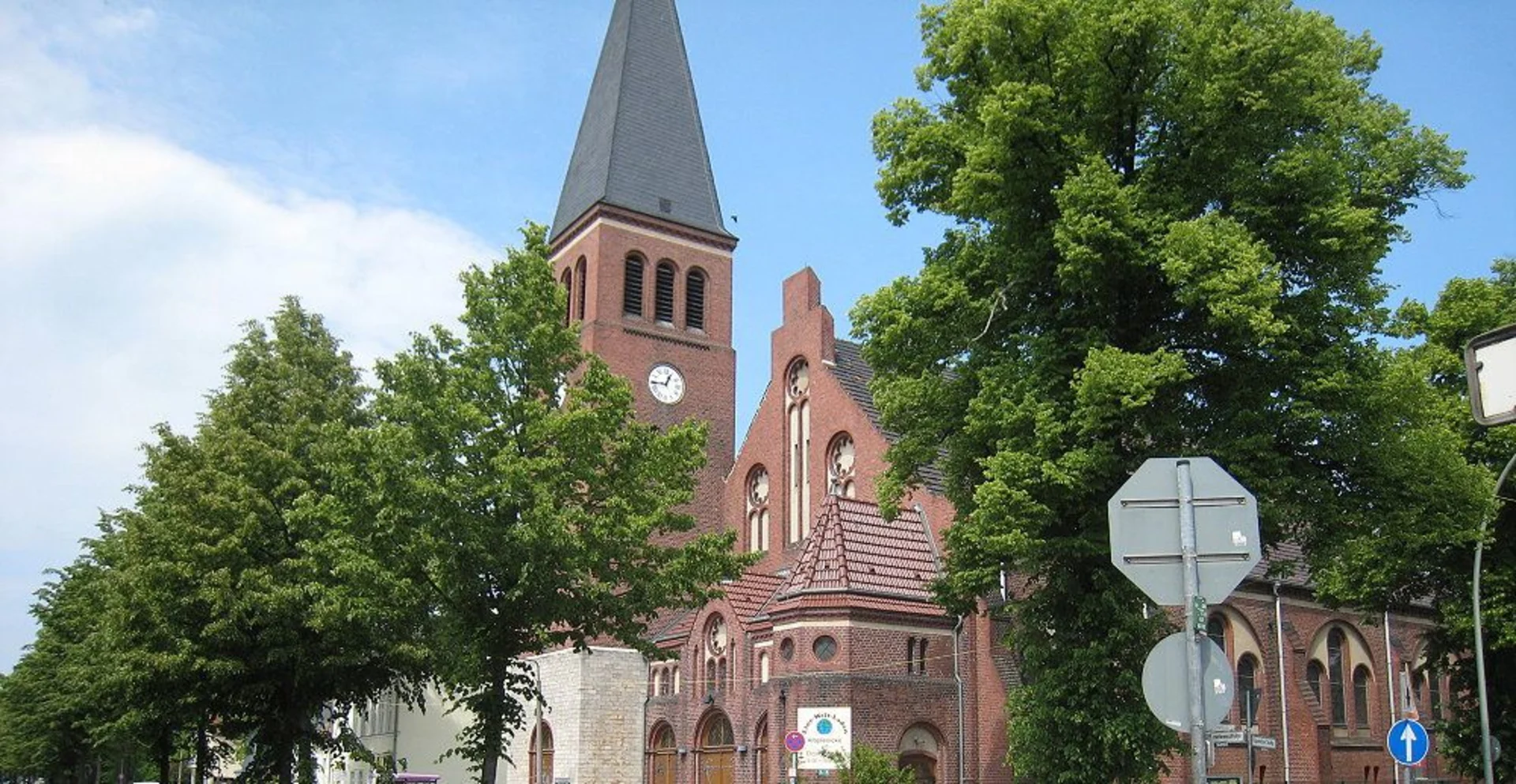 Backsteinkirche mit hohem Turm und Uhr, umgeben von grünen Bäumen und Straßenschildern.