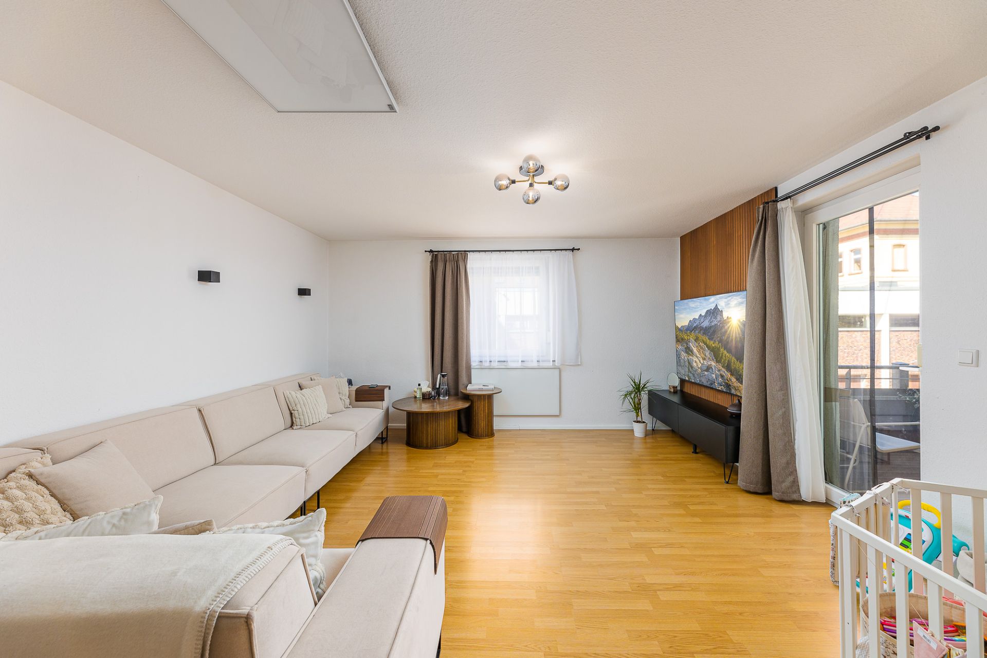 Helles Wohnzimmer mit Ecksofa, Fernseher, Holzboden und Fenster mit Vorhängen.