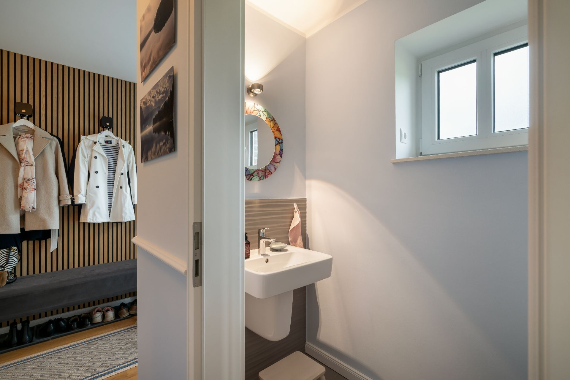 Badezimmer mit Waschbecken, rundem Spiegel und Fenster, daneben Flur mit Kleiderhaken und Schuhen.