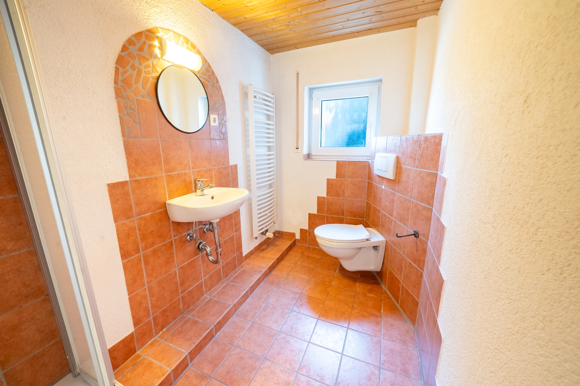 Helles Badezimmer mit Waschbecken, rundem Spiegel, Toilette, Fenster und orangefarbenen Fliesen.
