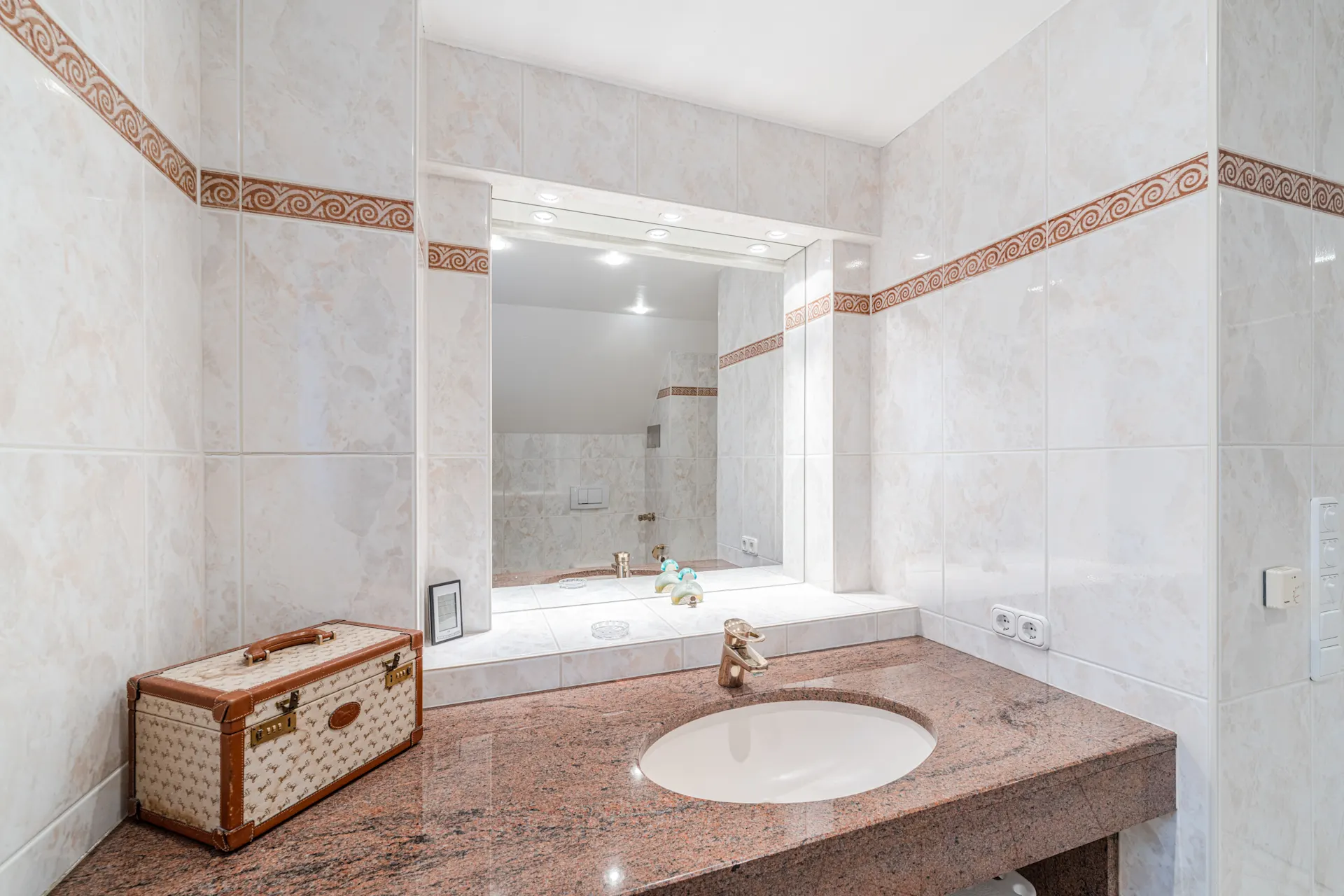 Badezimmer mit großem Spiegel, Marmorwaschbecken und einem Koffer auf der Ablage.