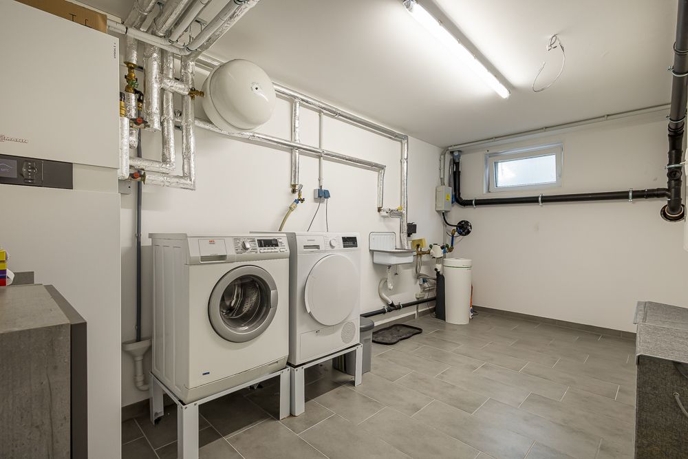 Waschraum mit Waschmaschine, Trockner, Waschbecken und technischen Anlagen, gefliestem Boden und kleinem Fenster.