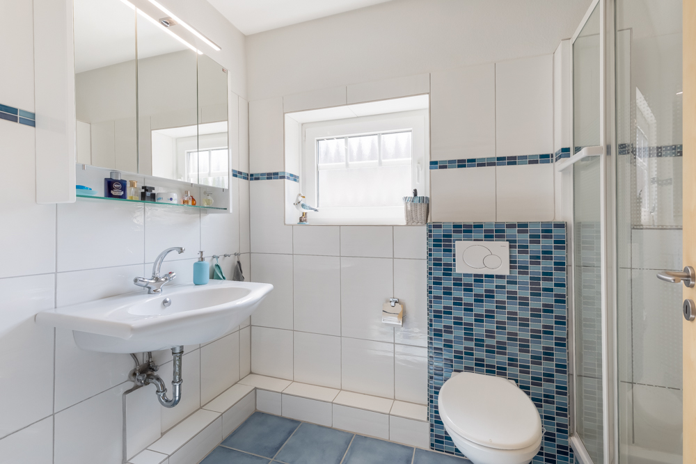 Modernes Badezimmer mit Waschbecken, Spiegel, Toilette, Fenster und Dusche mit blauen Fliesen.
