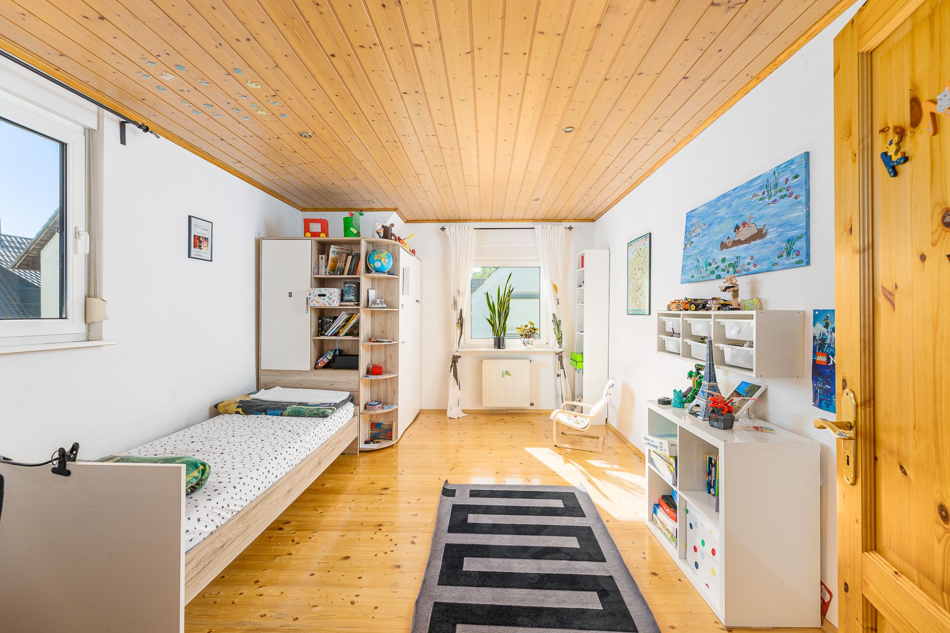 Helles Kinderzimmer mit Holzboden, Einzelbett, Regal, Teppich und großem Fenster.