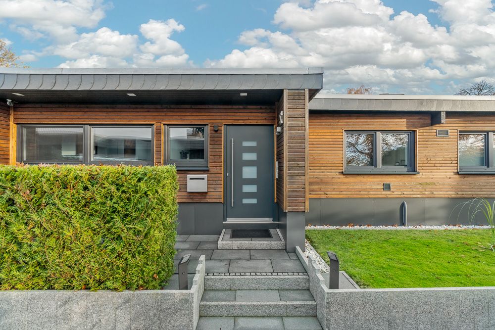 Moderne Hausfassade mit Holzelementen, grauer Tür und angrenzendem Garten.