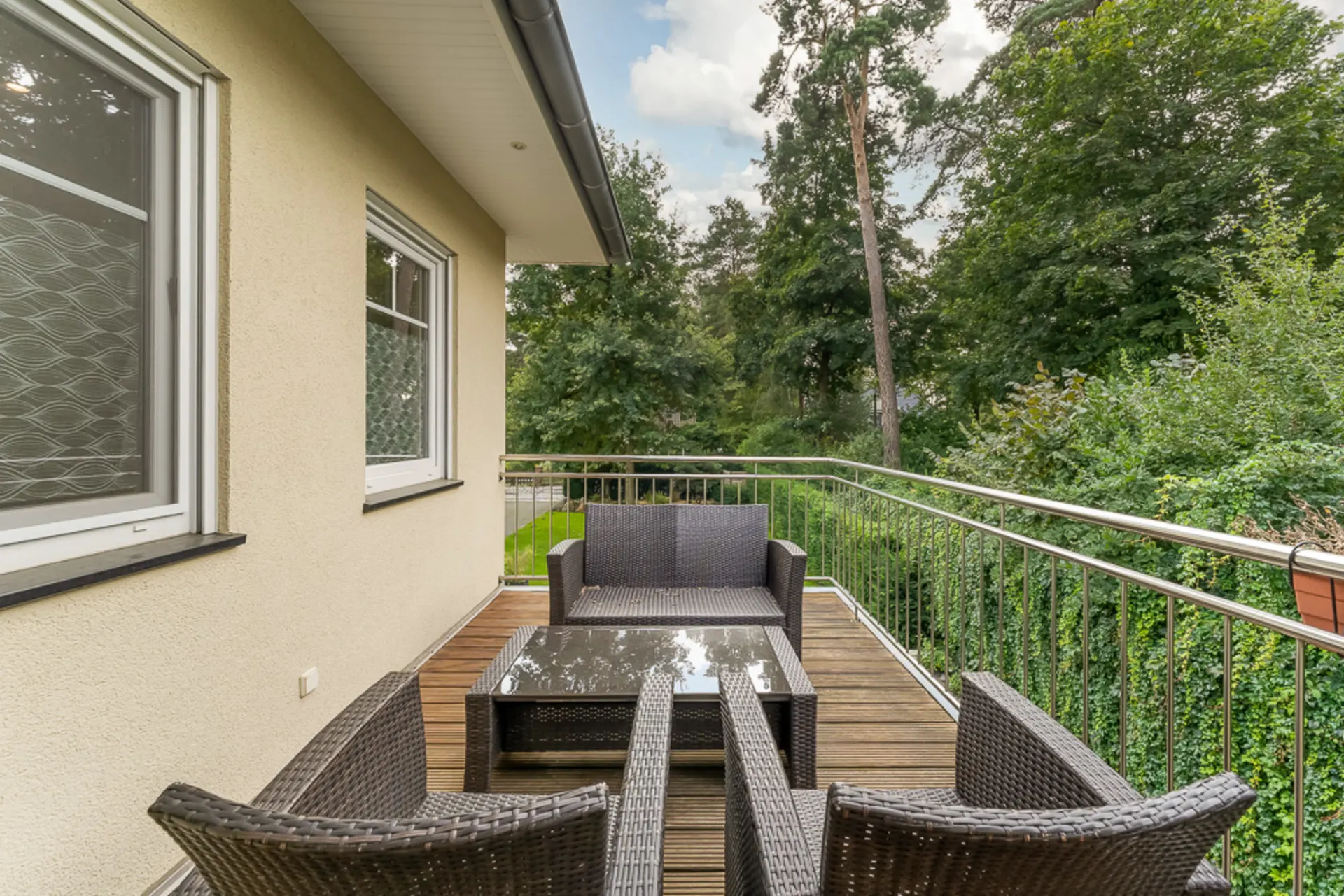 Balkon mit Rattanmöbeln und Glastisch, umgeben von Bäumen und Gartenblick.