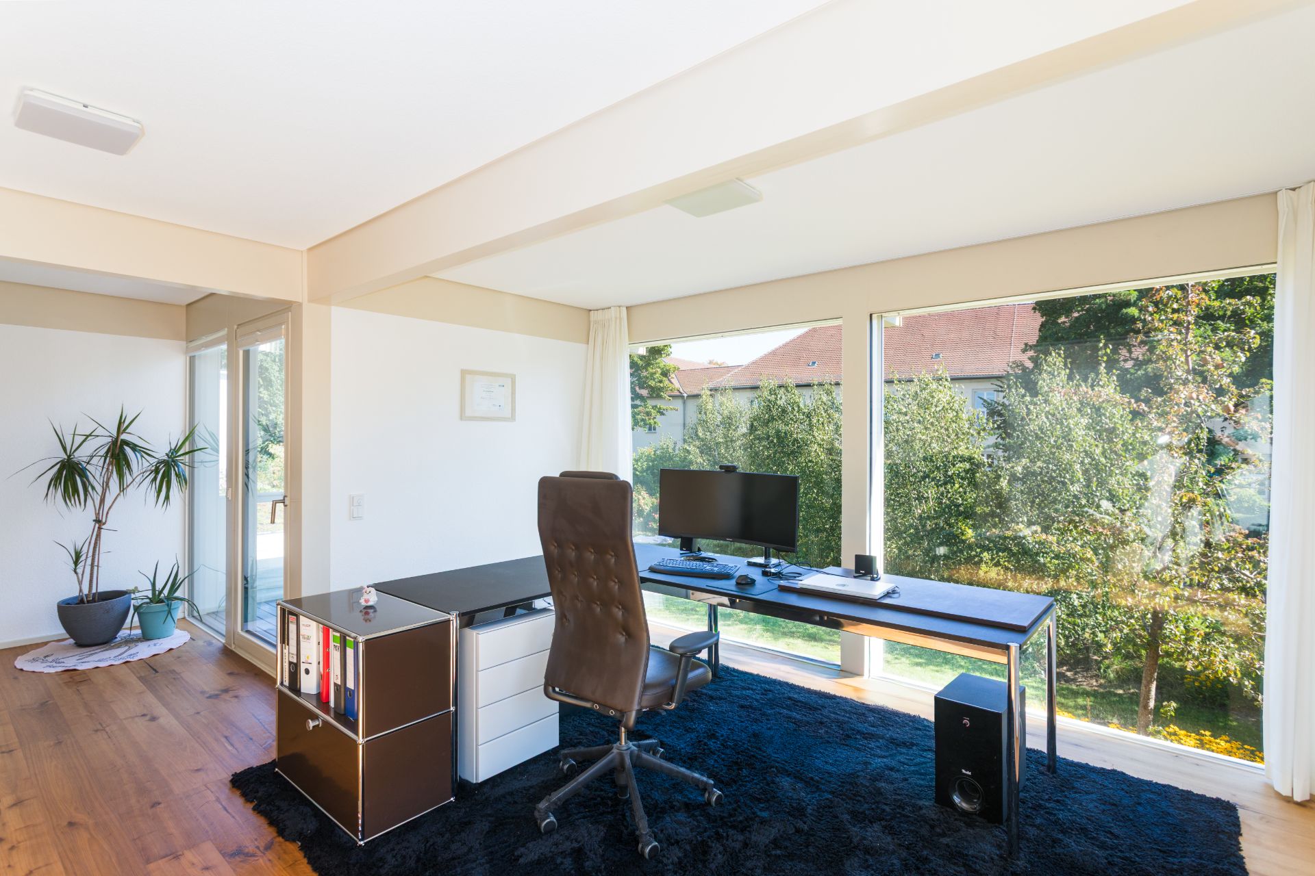 Modernes Arbeitszimmer mit großem Fenster, Schreibtisch, Stuhl und Computermonitor.