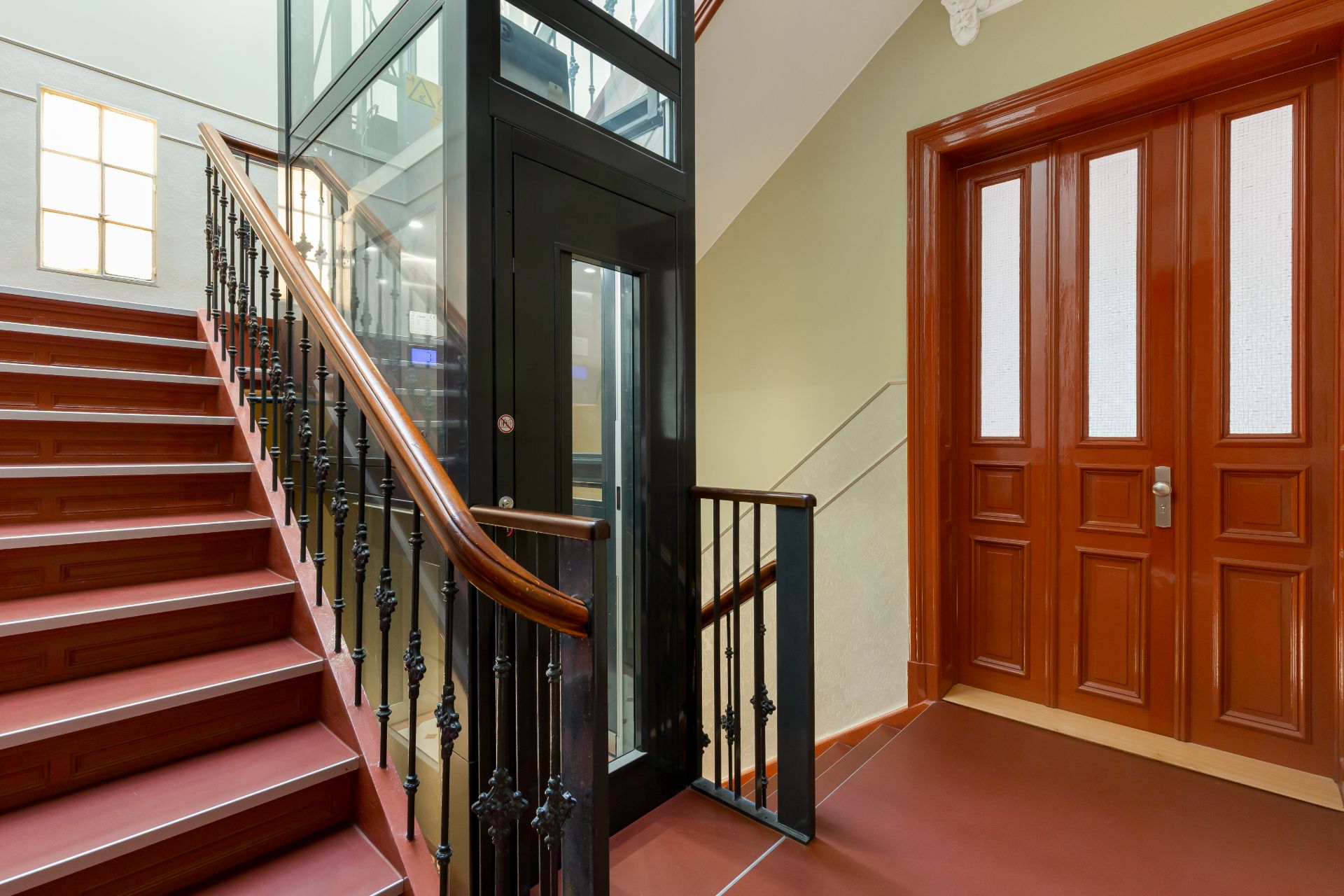 Treppe mit Holzgeländer, gläserner Aufzug und braune Eingangstür in einem Wohnhausflur.