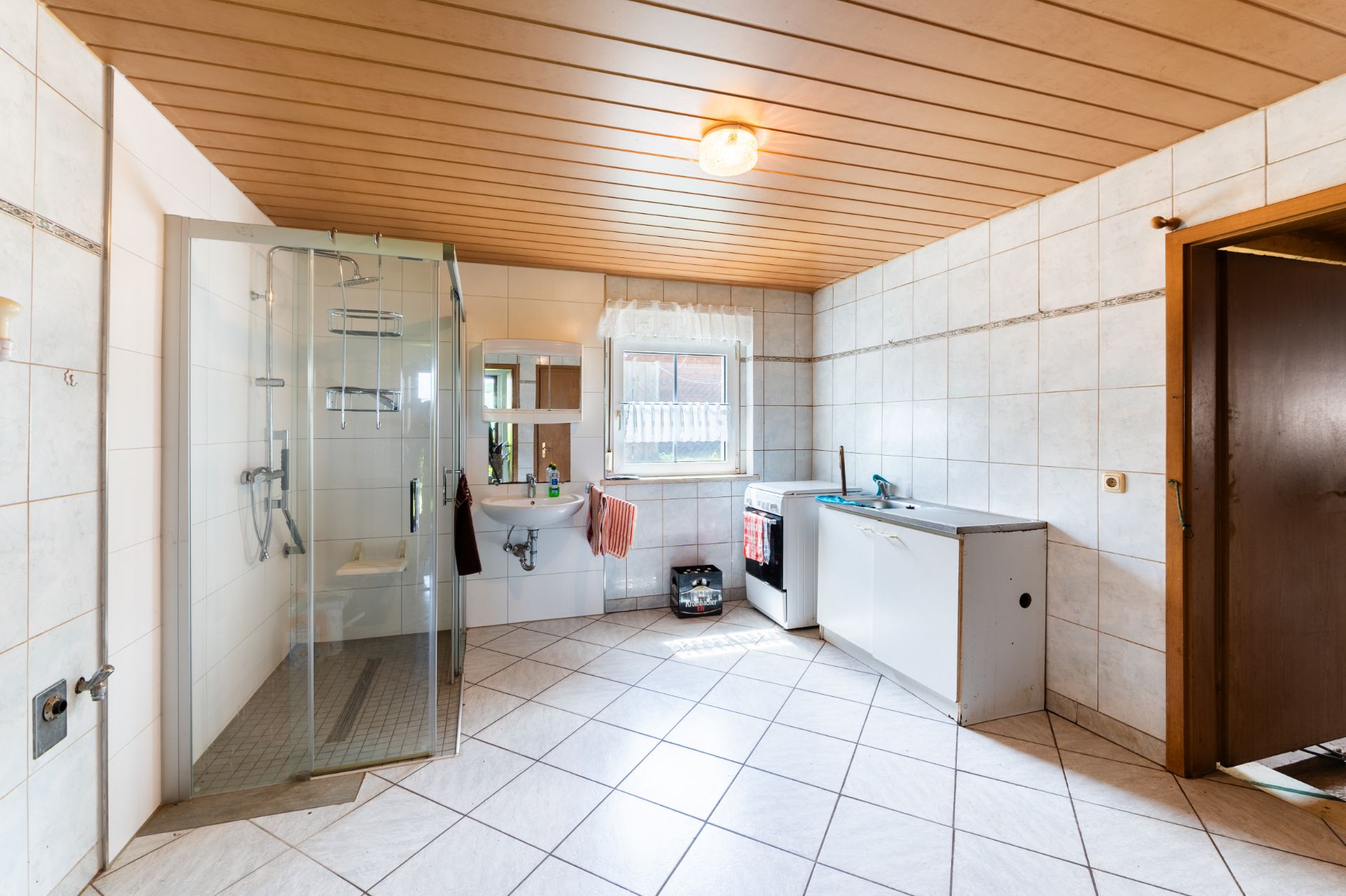 Helles Badezimmer mit Dusche, Waschbecken, Fenster und Waschmaschine auf gefliestem Boden.