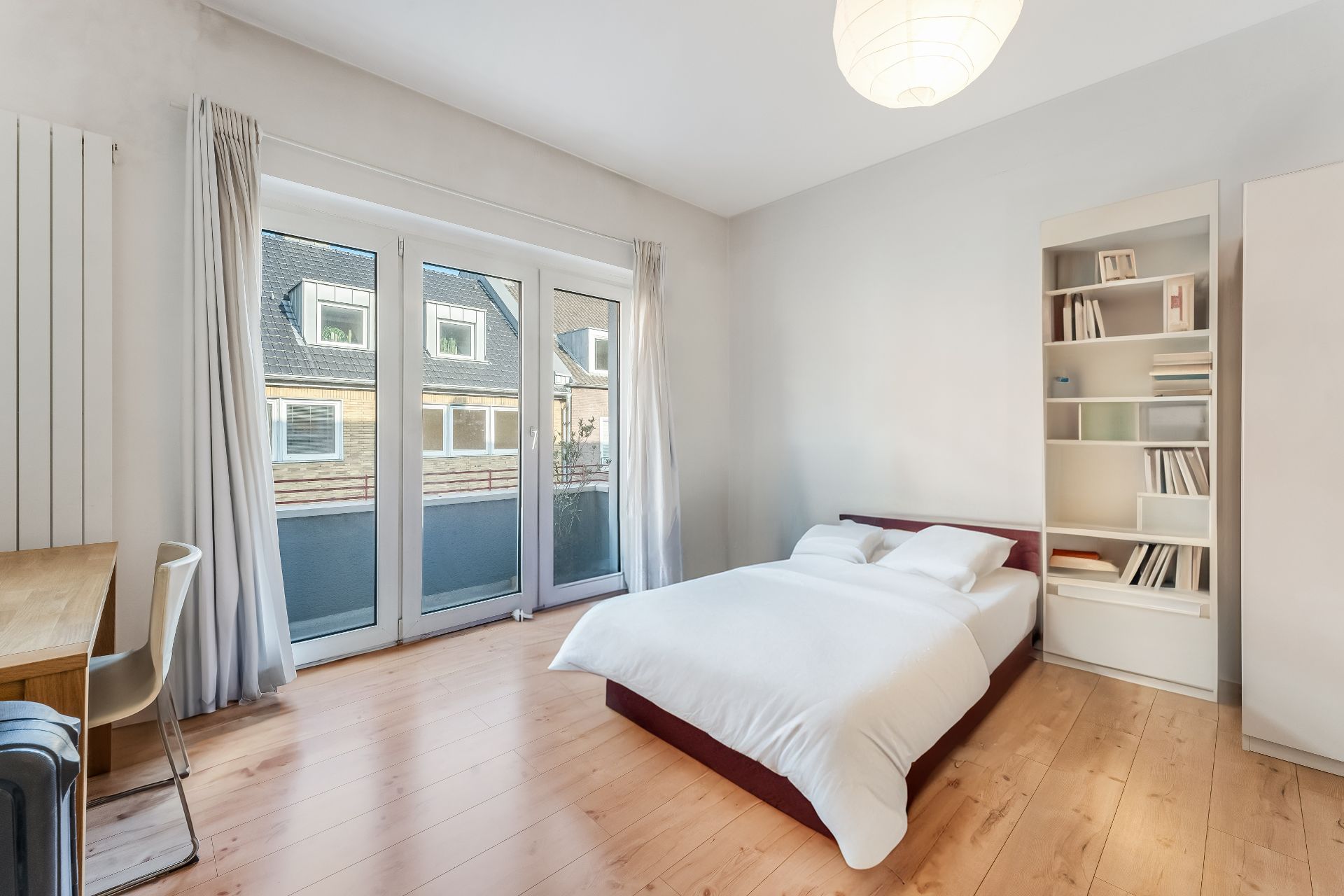 Helles Schlafzimmer mit Doppelbett, Schreibtisch, Bücherregal und großen Fenstern.