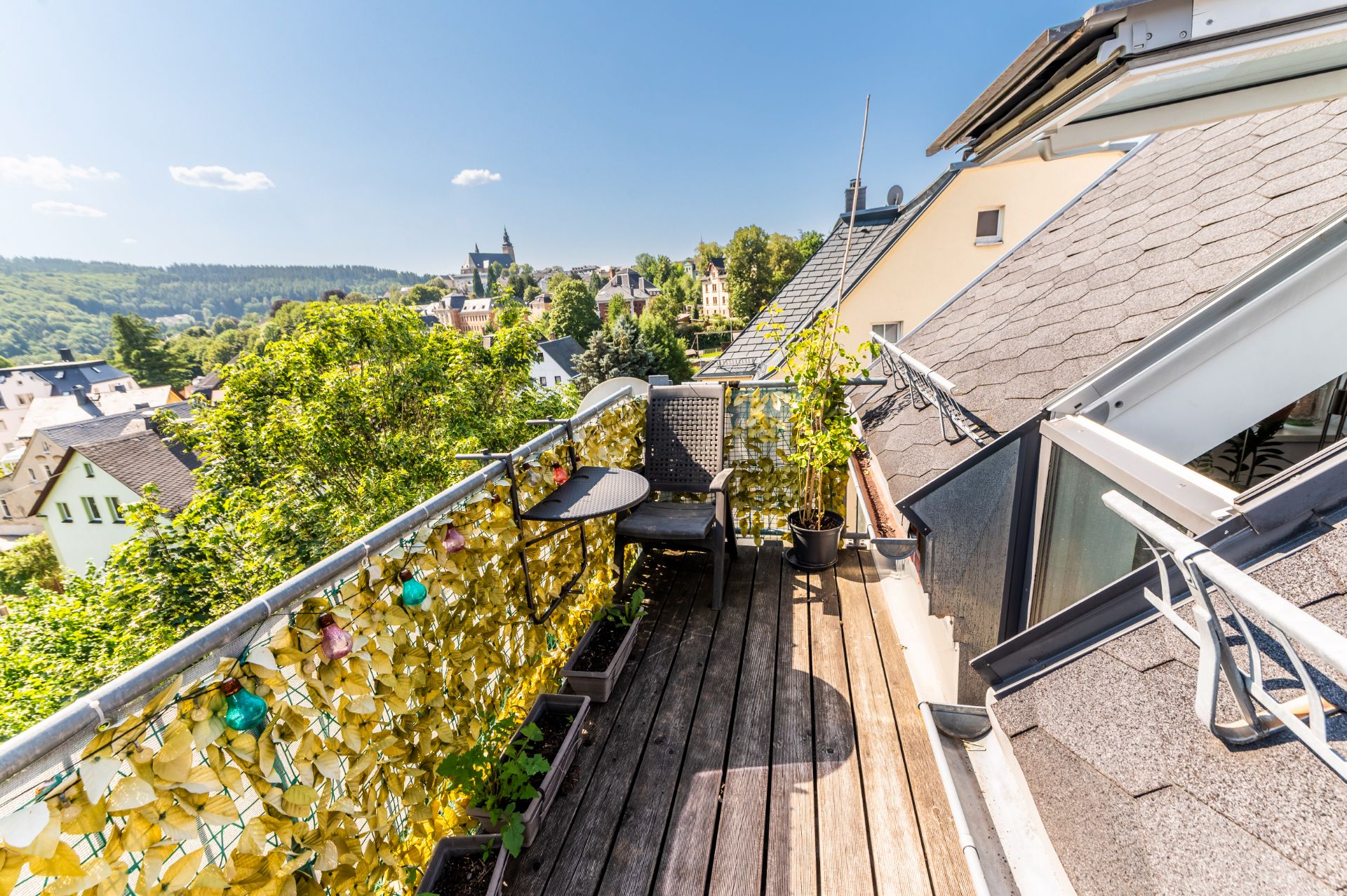 Sonnige Dachterrasse mit Holzfußboden, Pflanzen, Tisch und Stuhl, Blick auf grüne Landschaft und Häuser.