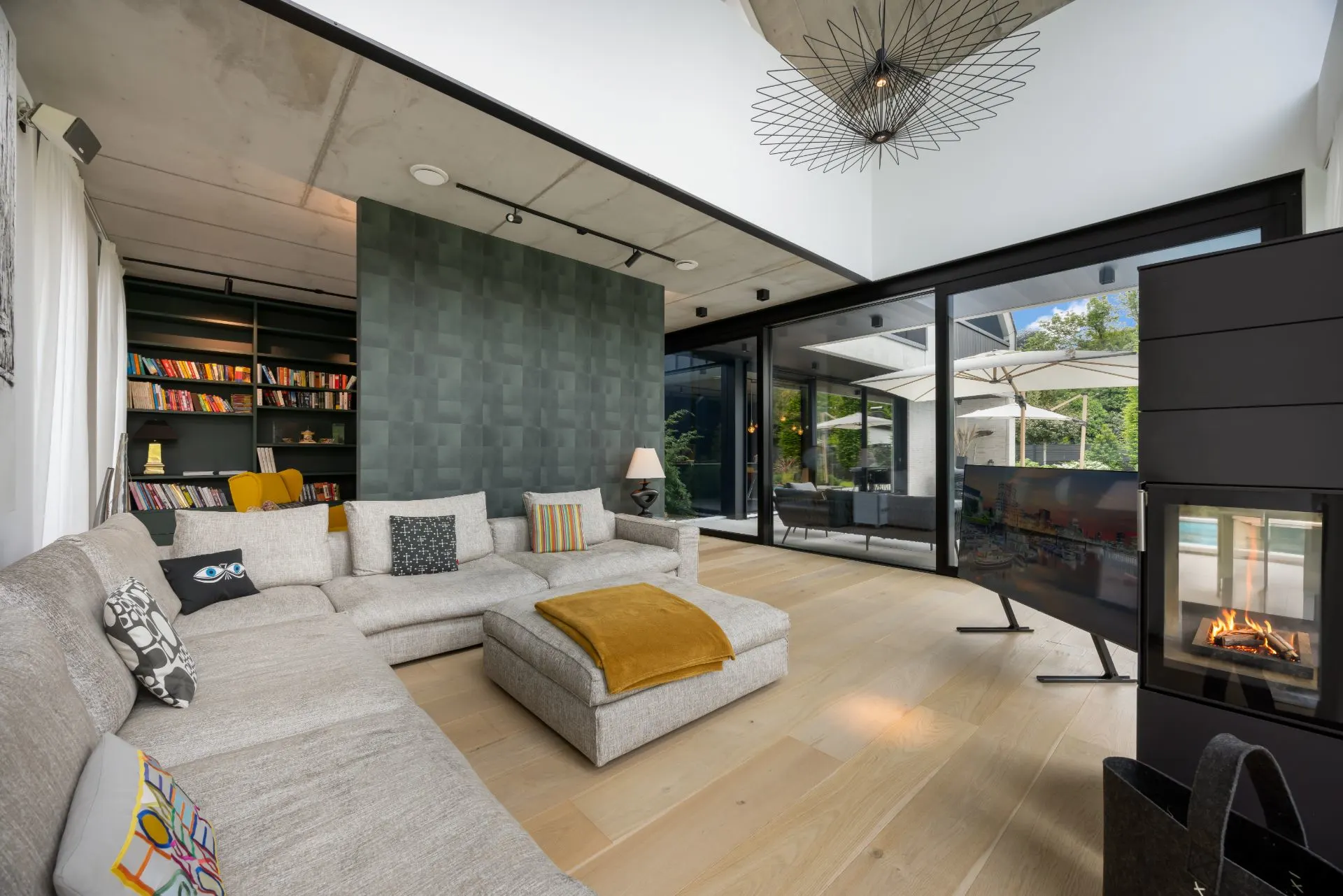 Modernes Wohnzimmer mit Ecksofa, Kamin, Bücherregal und großen Fenstern zu Terrasse und Garten.