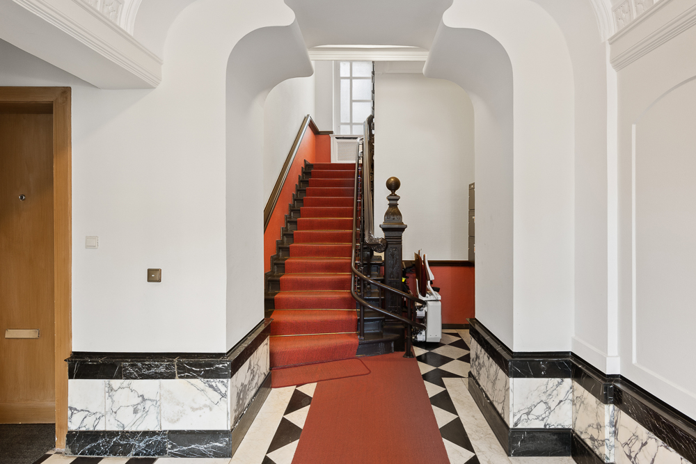Heller Flur mit Marmorboden und rotem Teppich, Treppe mit rotem Läufer nach oben.