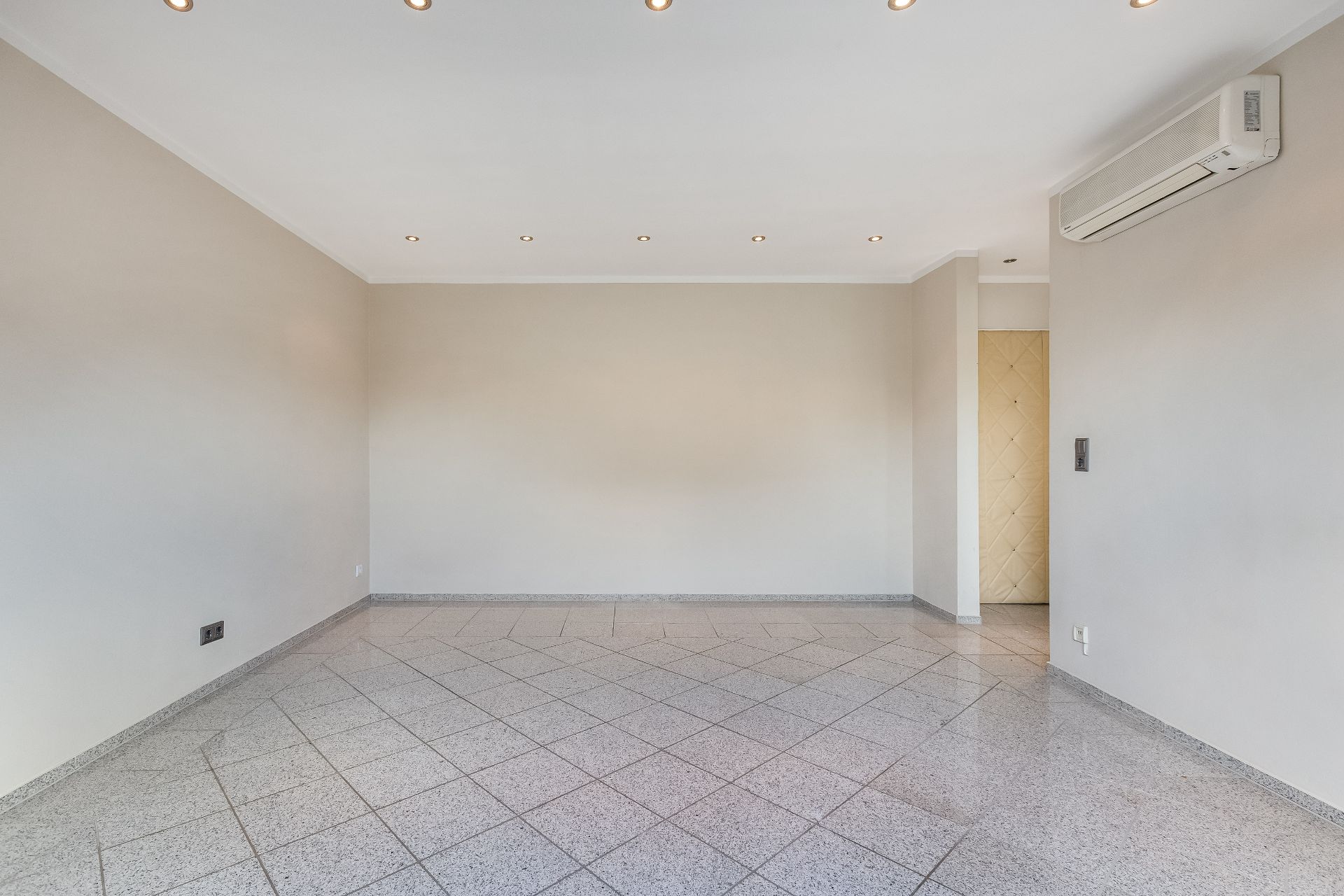 Leerer Raum mit Fliesenboden, weißen Wänden und Deckenleuchten, Klimaanlage an der Wand.