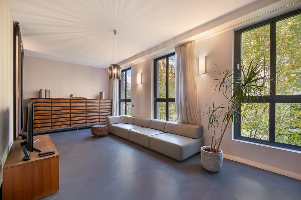 Modernes Wohnzimmer mit großer Eckcouch, Holzkommode, TV, großen Fenstern und Zimmerpflanze.