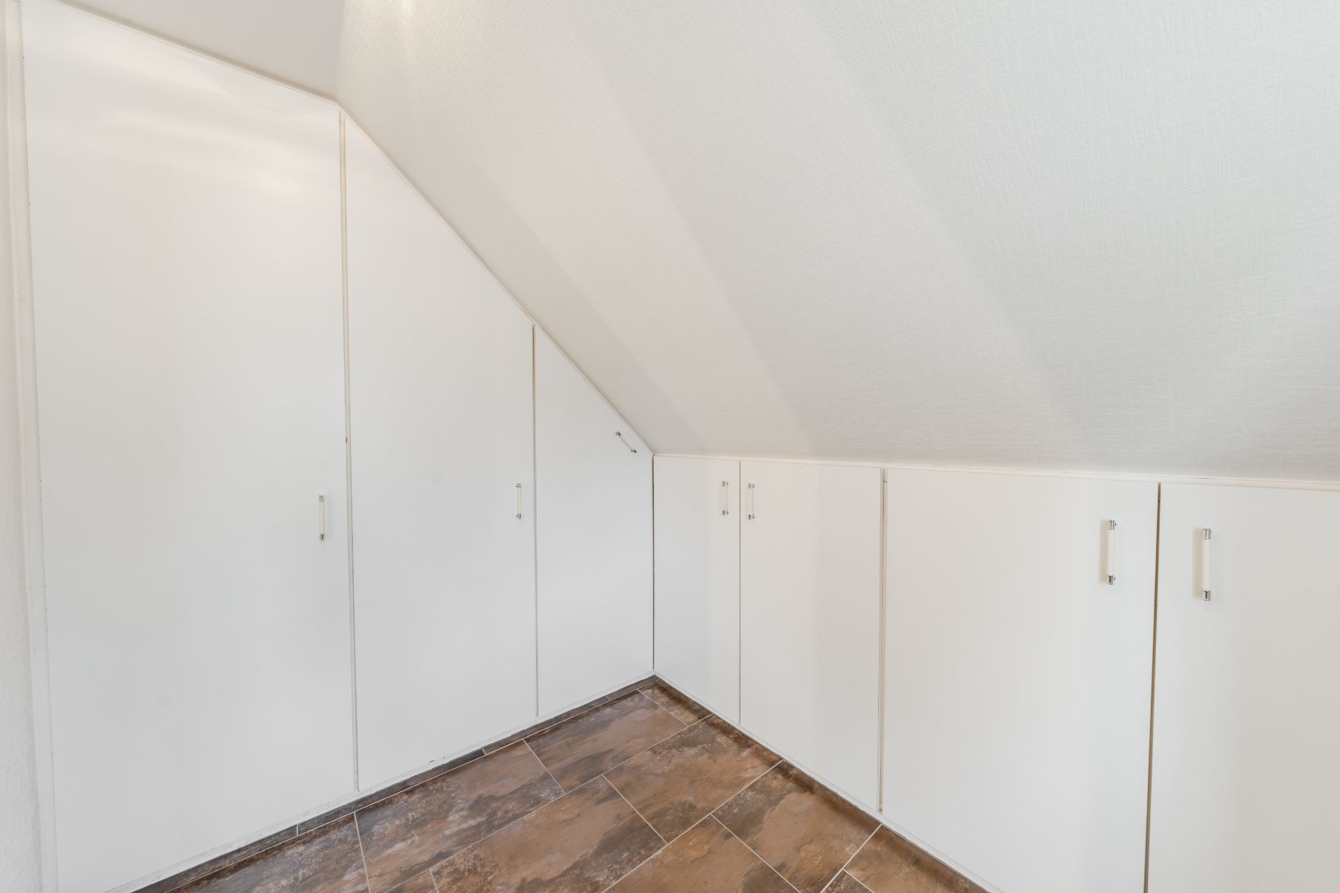 Raum mit eingebauten weißen Schränken unter einer Dachschräge und braunem Fliesenboden.