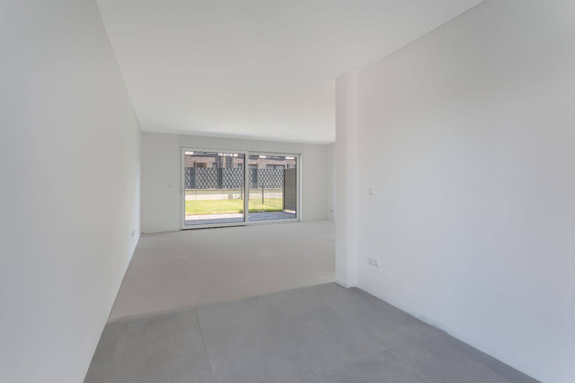 Helles, leeres Wohnzimmer mit großen Fenstern und Blick auf einen Garten.