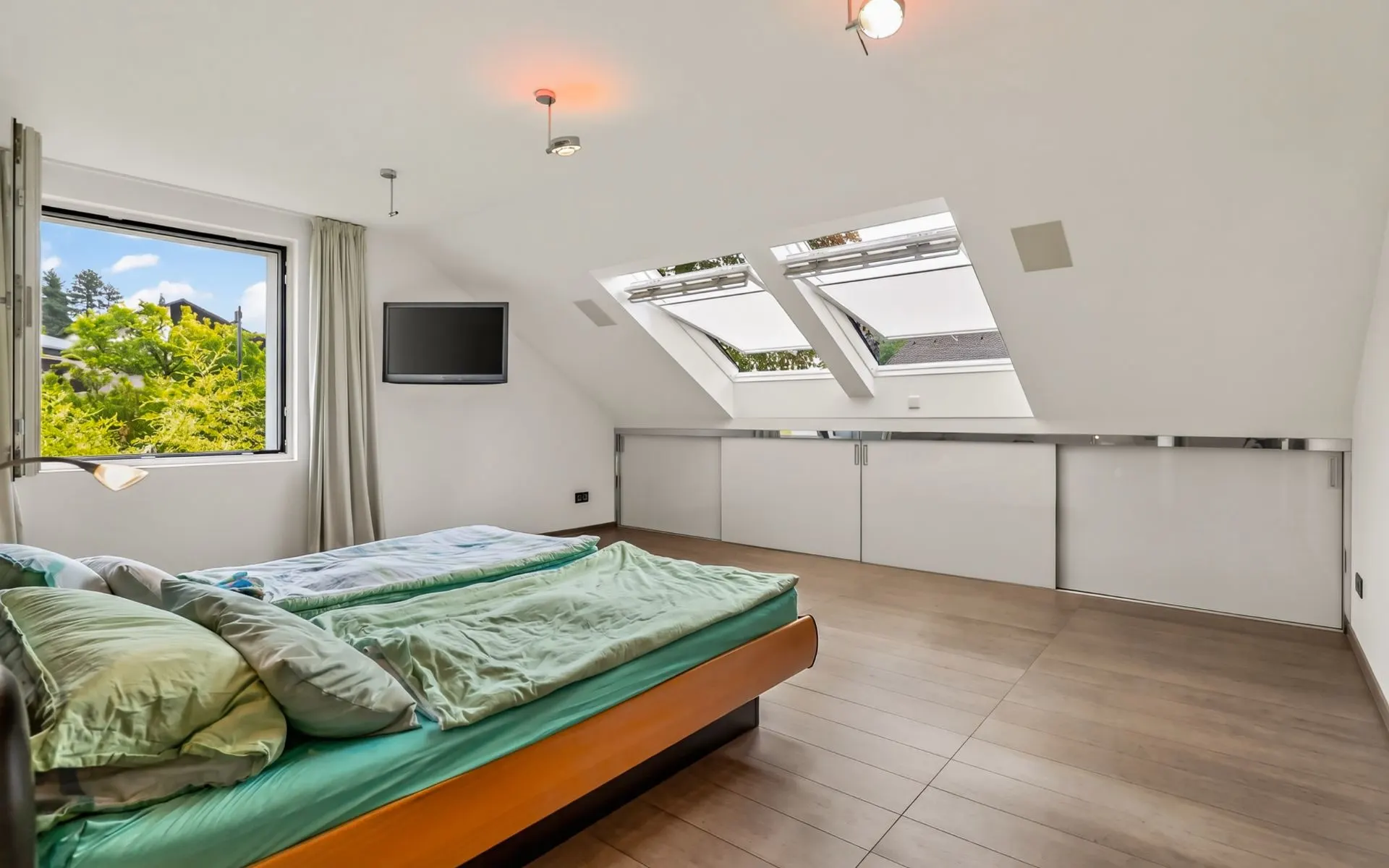 Modernes Schlafzimmer mit Dachfenstern, Doppelbett, Holzfußboden und Fenster mit Blick ins Grüne.