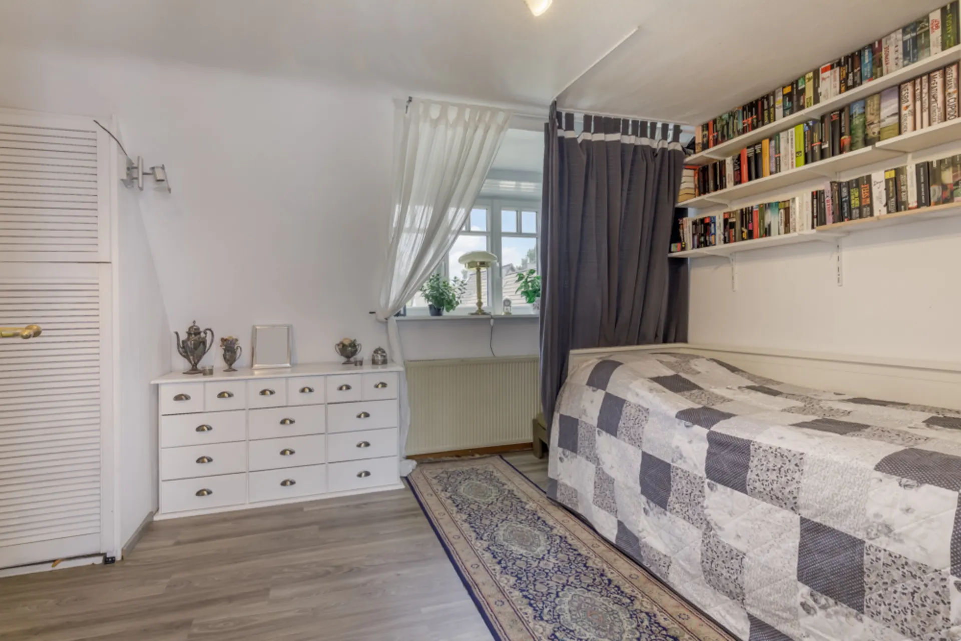 Helles Schlafzimmer mit Einzelbett, Bücherregalen, Kommode und Fenster mit Vorhängen.