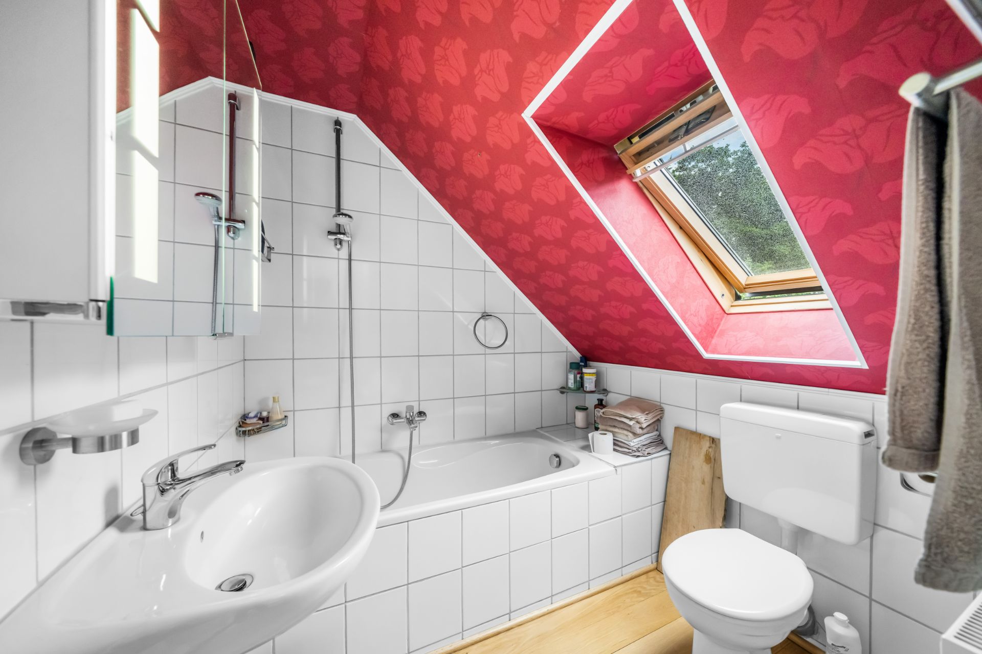 Kleines Badezimmer mit Badewanne, Waschbecken, WC und Dachfenster unter roter Schräge.