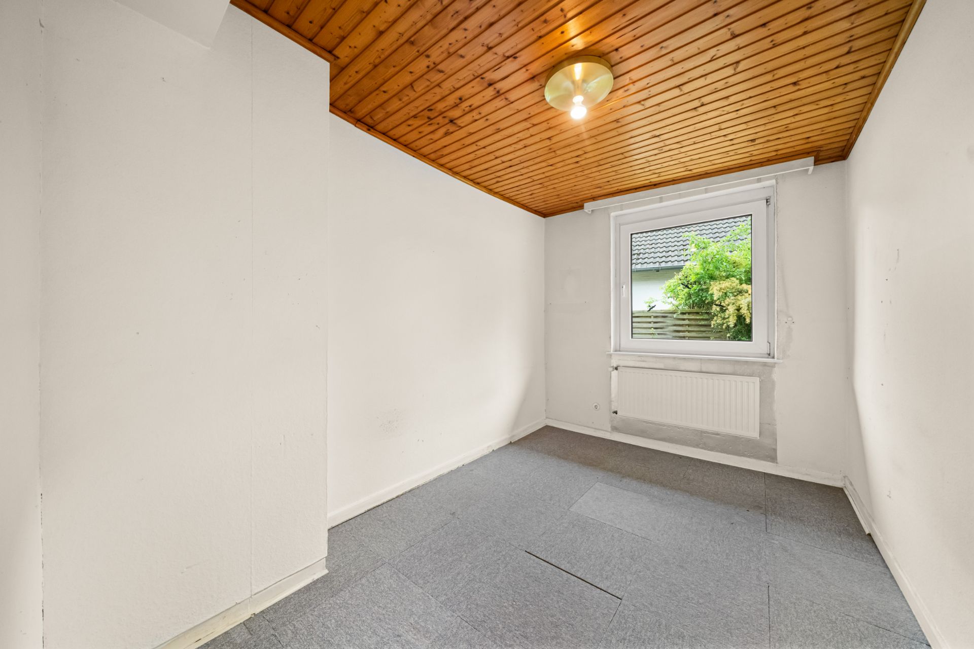 Kleines, leeres Zimmer mit Holzdecke, weiß gestrichenen Wänden und einem Fenster mit Blick nach draußen.