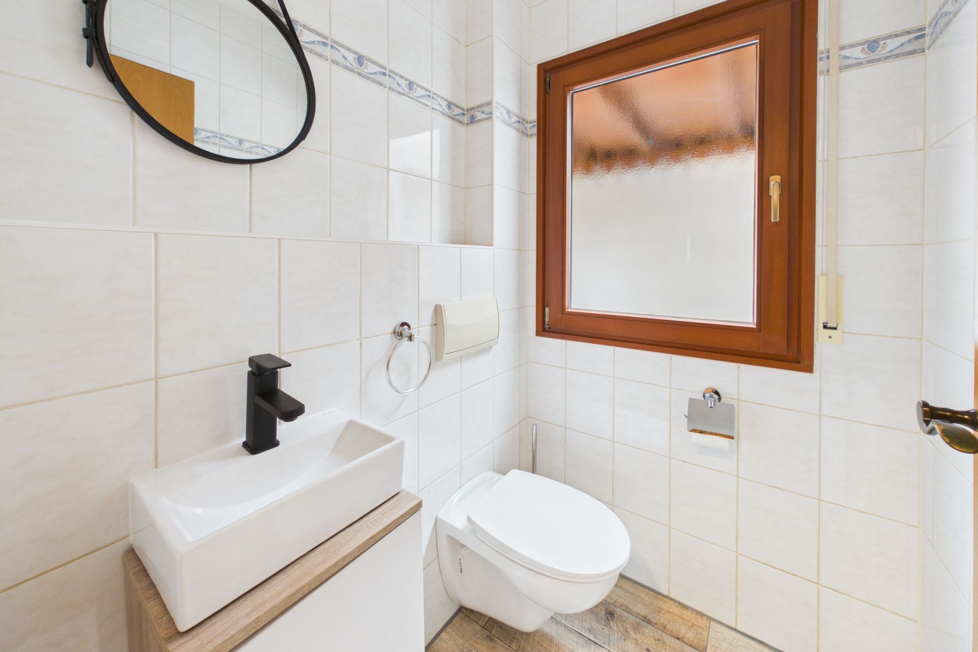 Helles Badezimmer mit Wandtoilette, Waschbecken, rundem Spiegel und großem Fenster.