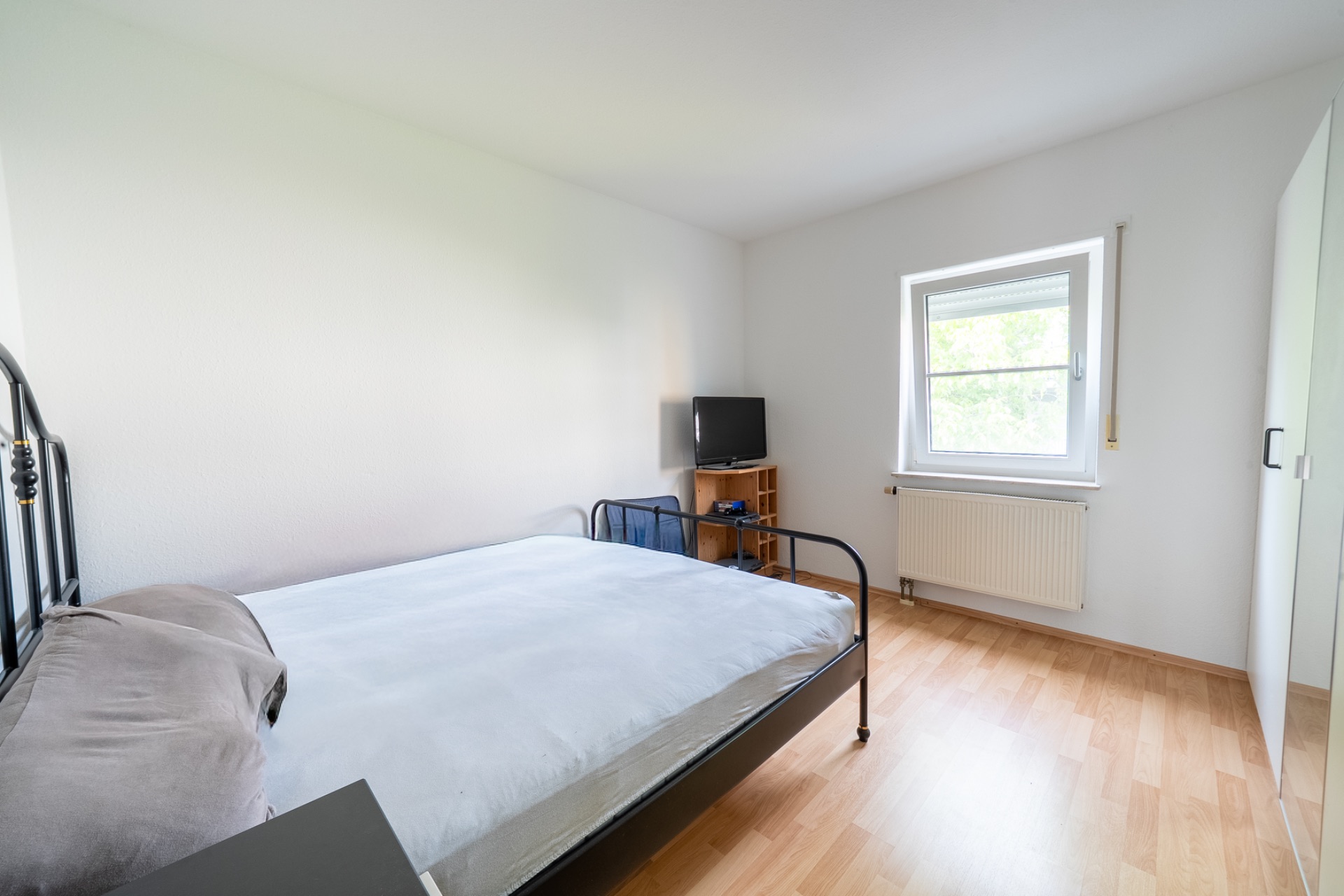 Helles Schlafzimmer mit Metallbett, kleinem Fernseher, Fenster und Kleiderschrank auf Holzboden.