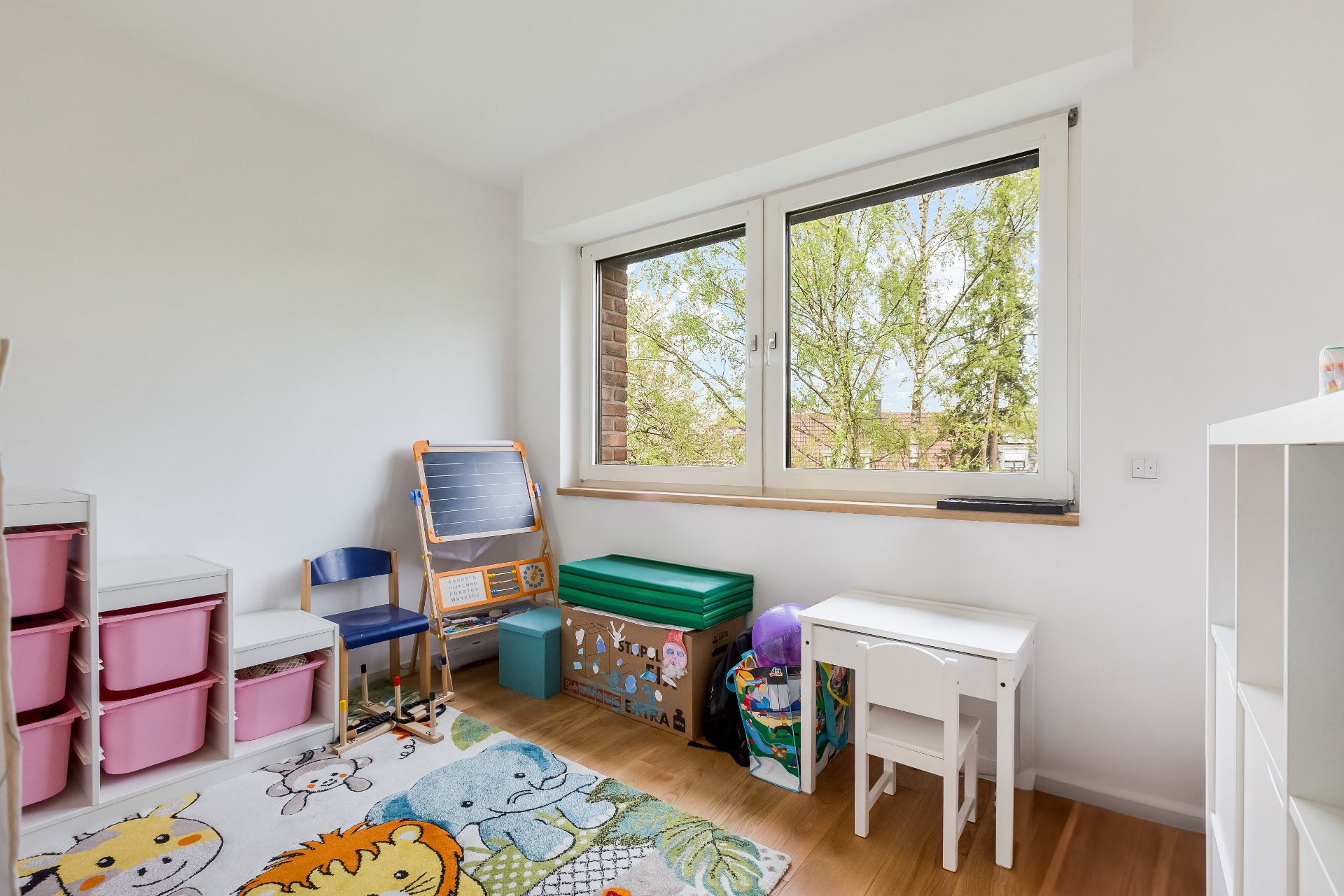 Helles Kinderzimmer mit Spielzeug, buntem Teppich, Kindertisch und großem Fenster.