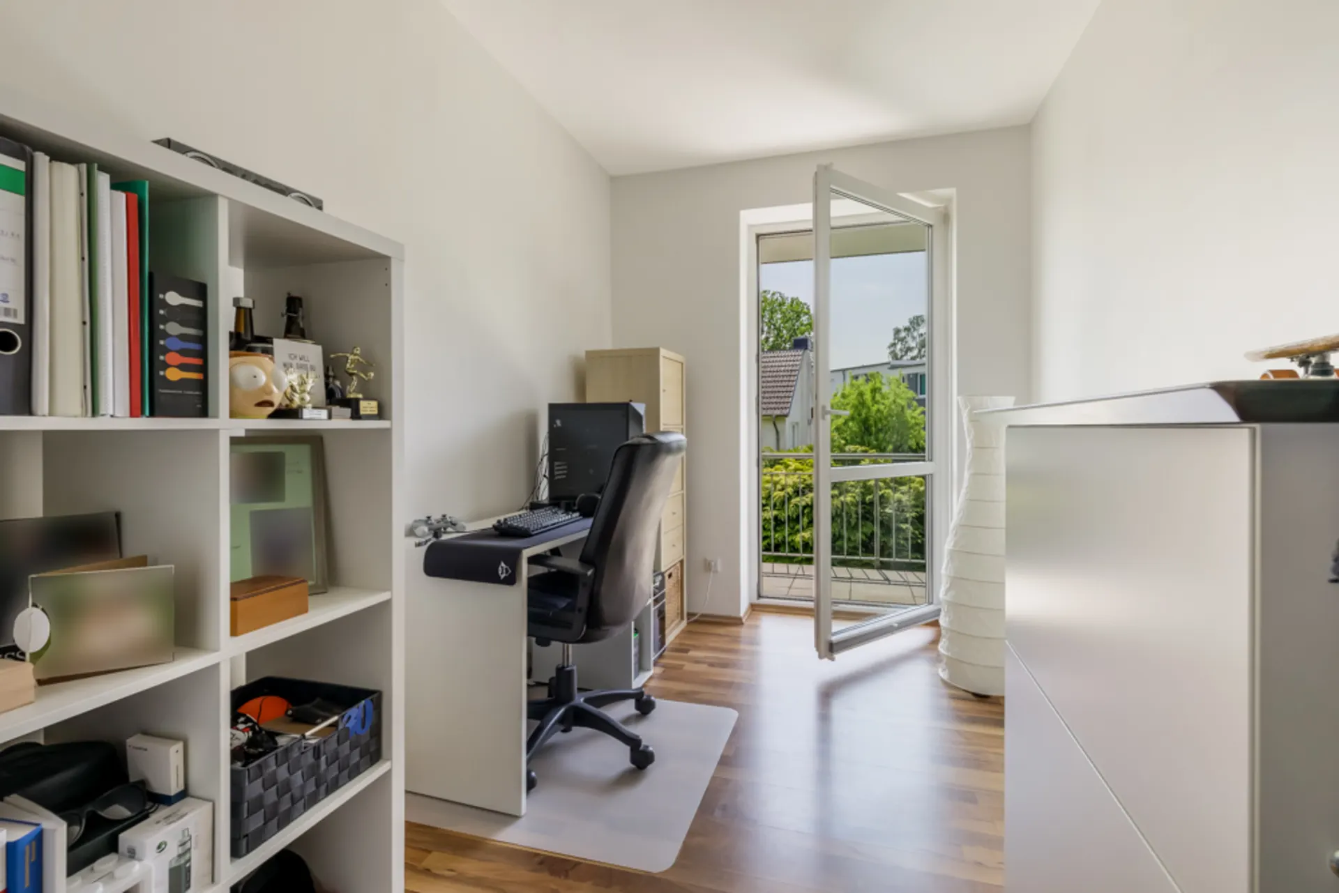 Helles Arbeitszimmer mit Schreibtisch, Regal und offenem Fenster zum Balkon.