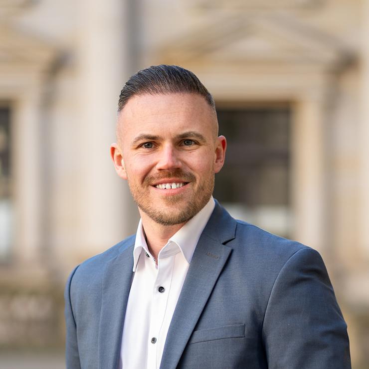 Ein lächelnder Mann im Anzug steht draußen vor einem unscharfen Gebäude.