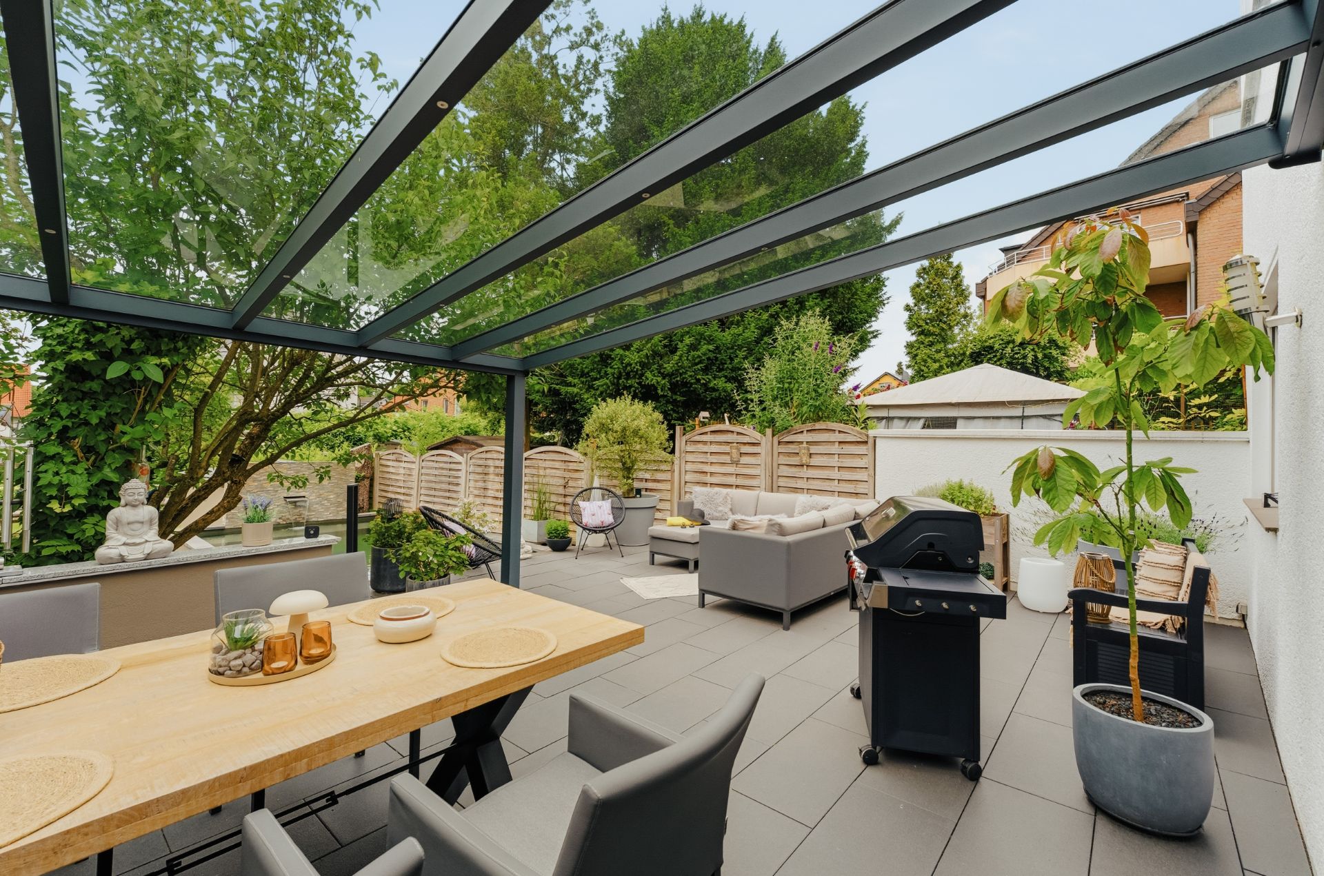 Überdachte Terrasse mit Esstisch, Grill, Pflanzen und Sitzecke im Garten eines Wohnhauses.
