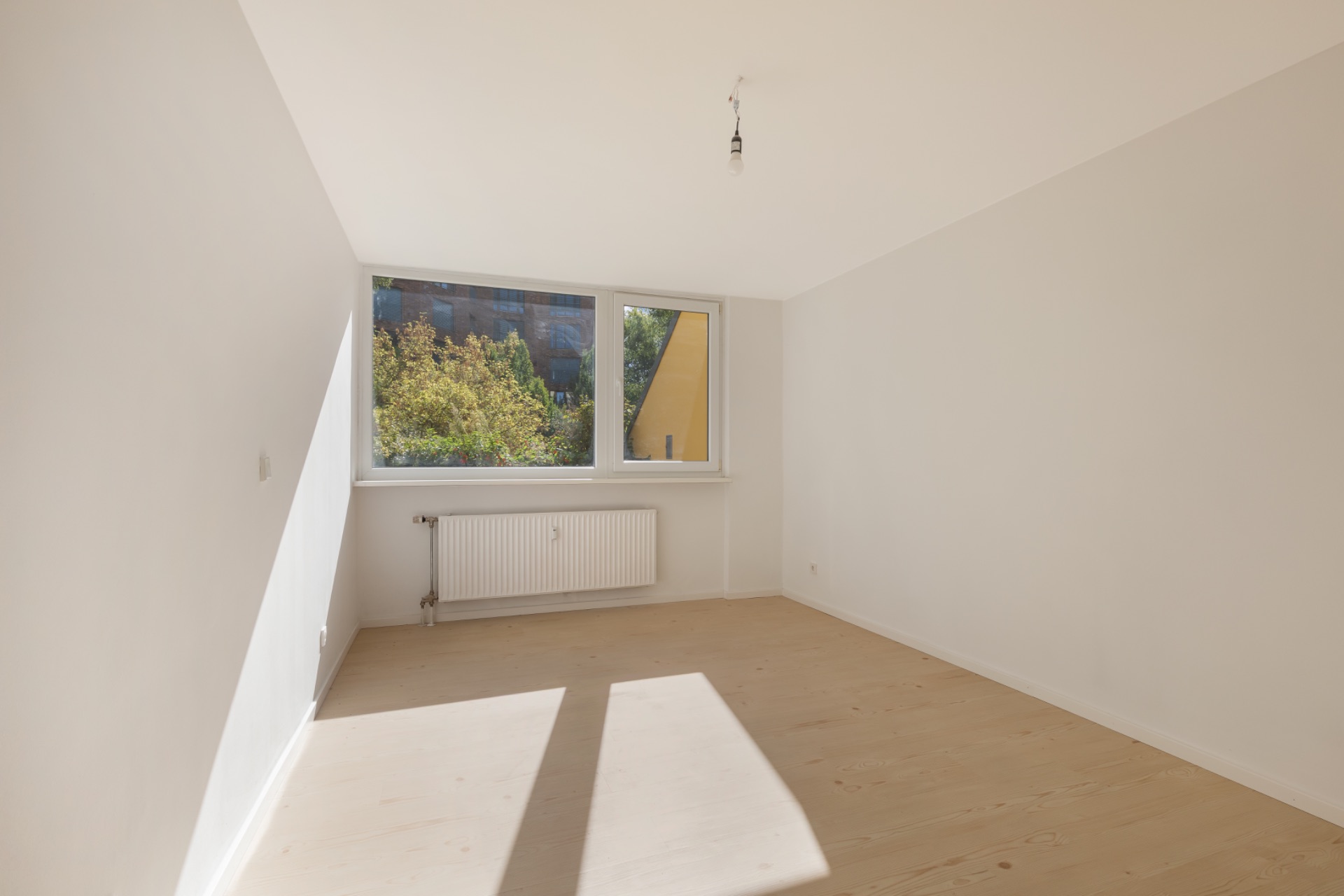 Helles, leeres Zimmer mit großem Fenster, Holzboden und Heizkörper unter dem Fenster.