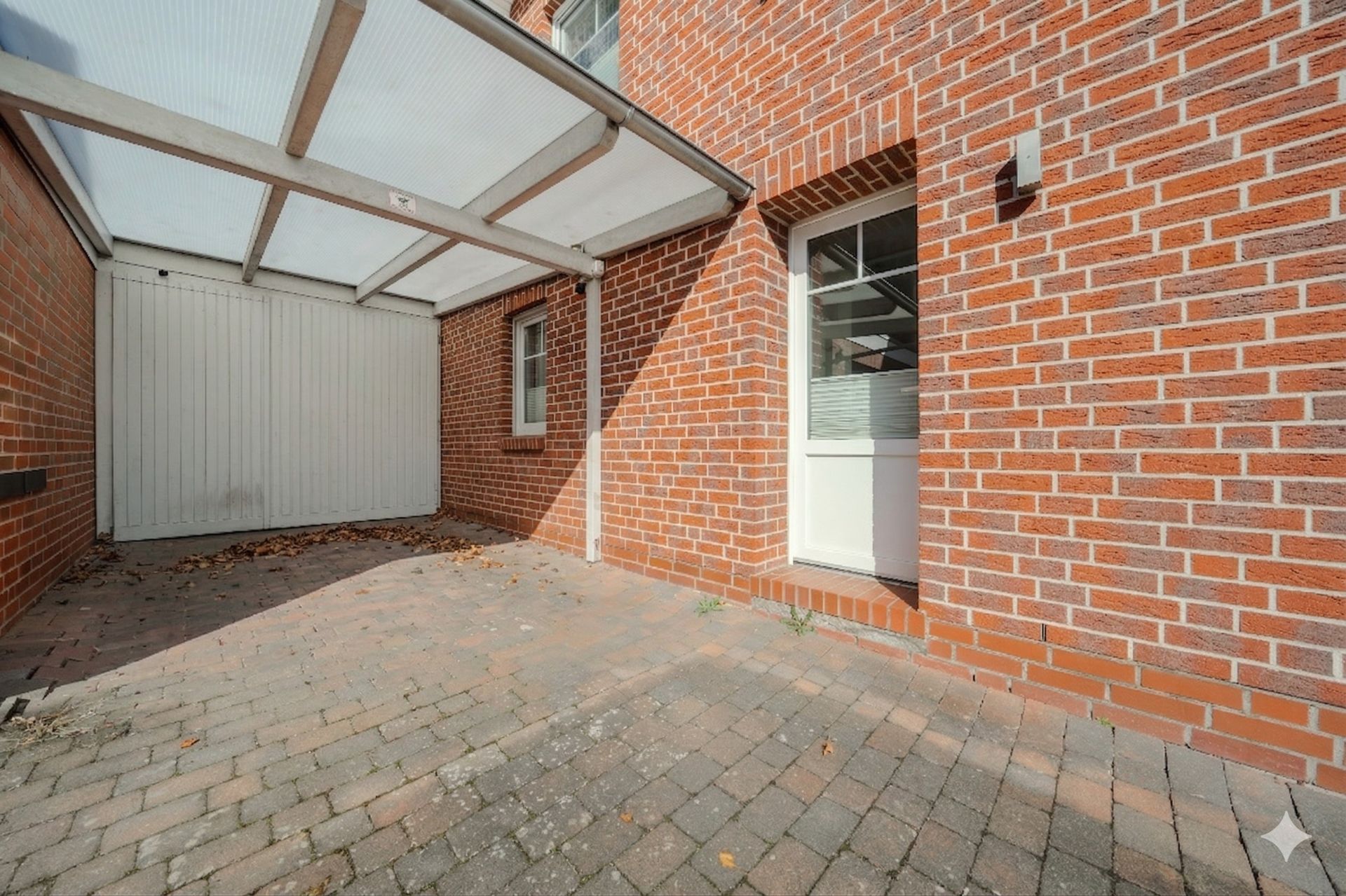 Überdachter Bereich mit Pflastersteinen und roter Backsteinfassade, weiße Tür und Fenster.