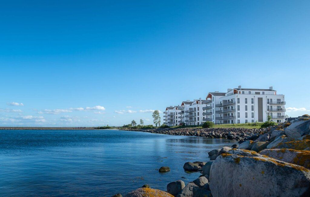 Weiße Wohngebäude am Wasser mit Steinen im Vordergrund und blauem Himmel.