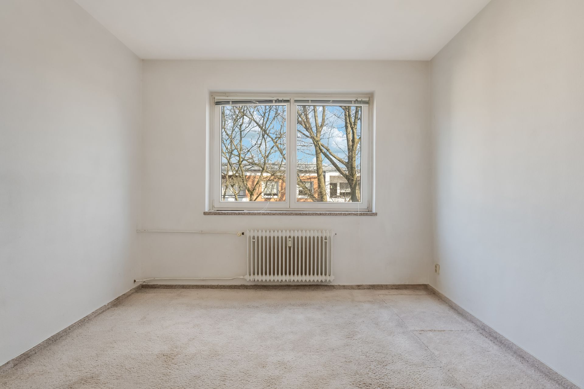 Leeres Zimmer mit Teppichboden, weiß gestrichenen Wänden und großem Fenster mit Blick auf Bäume.