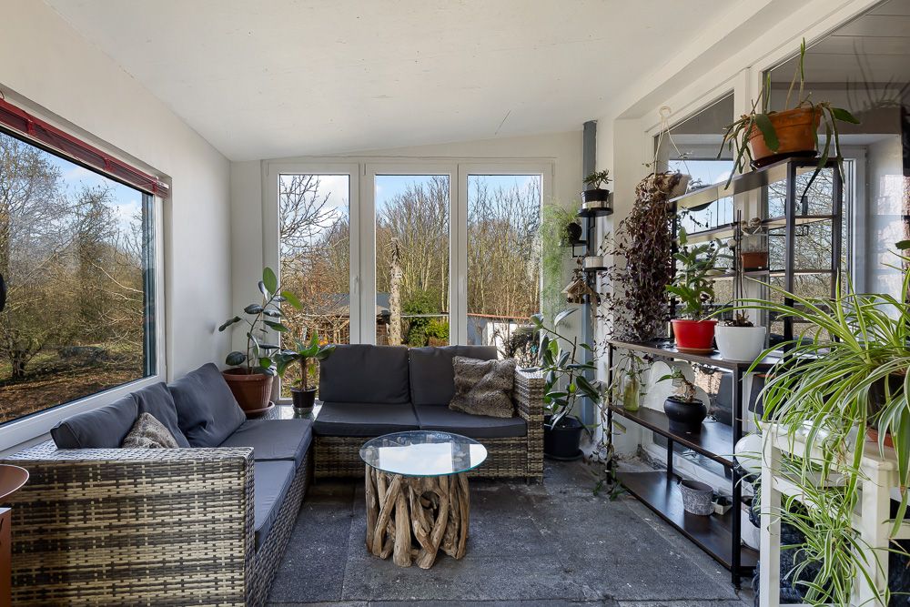 Wintergarten mit Ecksofa, Glastisch und vielen Zimmerpflanzen vor großen Fenstern.