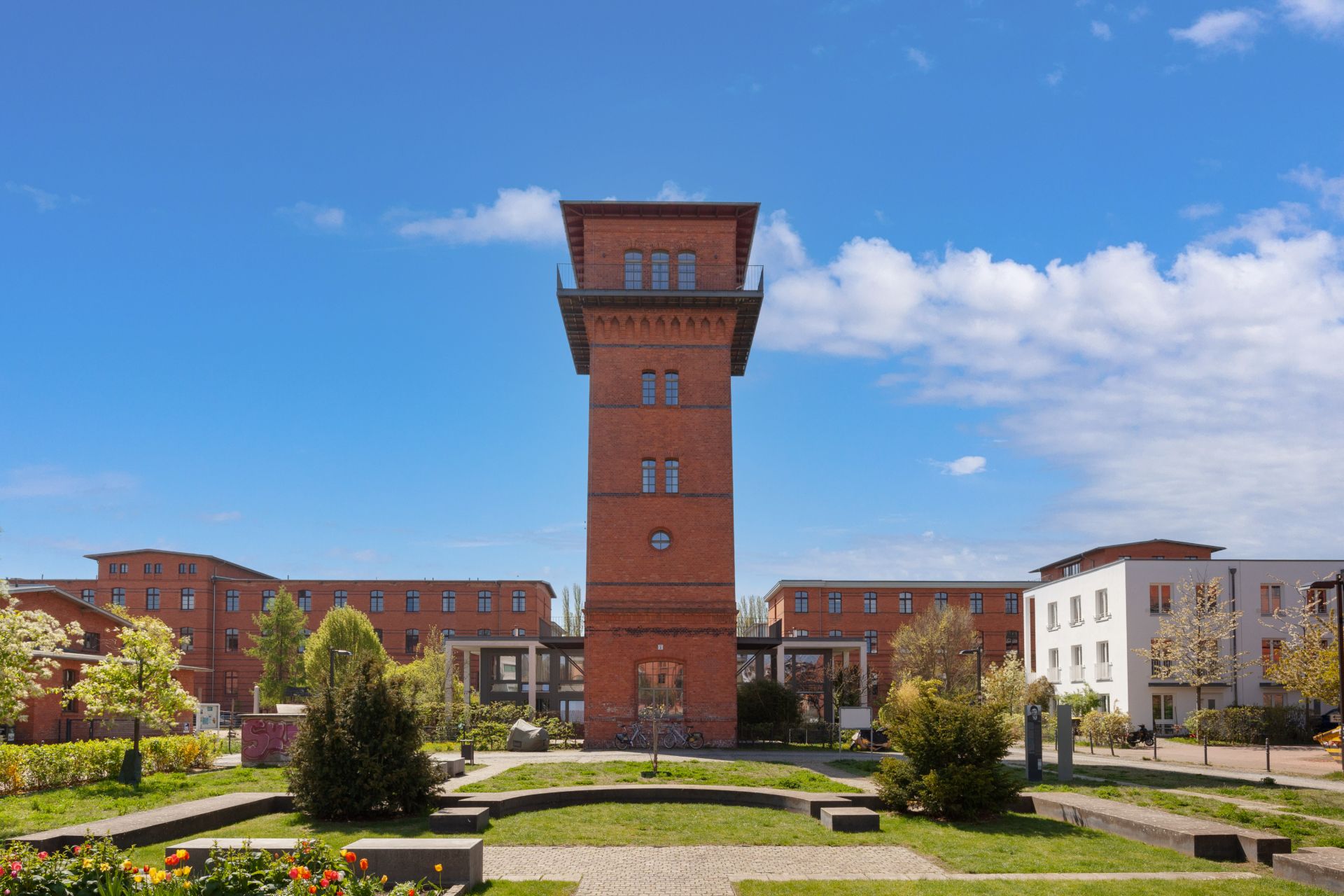 Roter Backsteinturm inmitten einer Wohnanlage mit Garten und Bänken im Vordergrund.