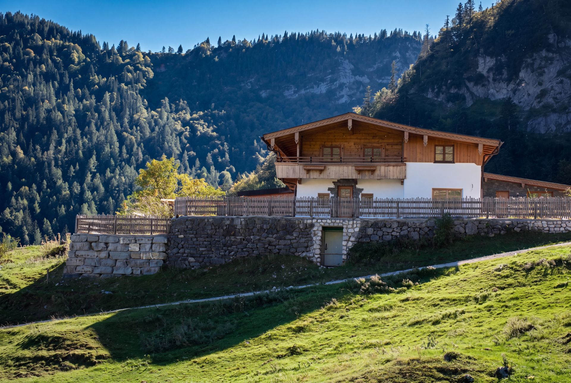 Haus im Alpenstil auf einem Hügel, umgeben von Bergen und Wiese, Holzzaun im Vordergrund.