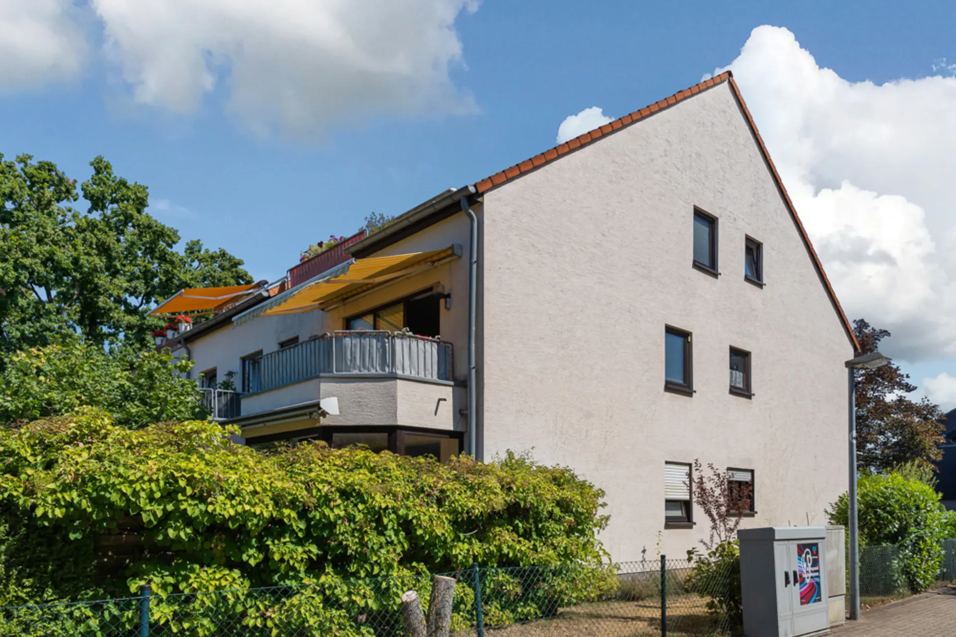 Zweistöckiges Wohnhaus mit Balkon und gelben Markisen, umgeben von grünen Büschen.