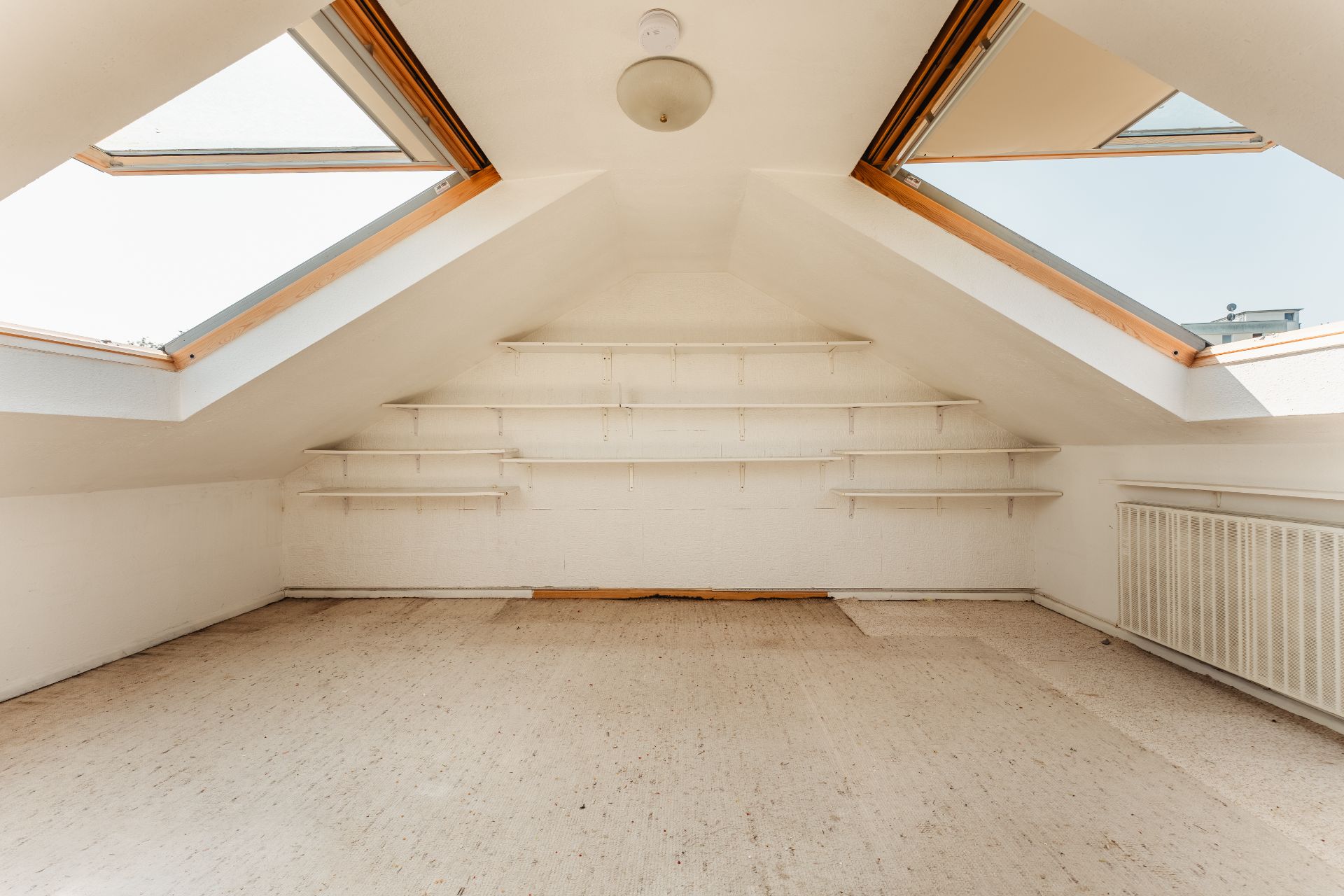 Leerer Dachraum mit Schrägfenstern, Regalbrettern an der Wand und Heizkörper rechts.