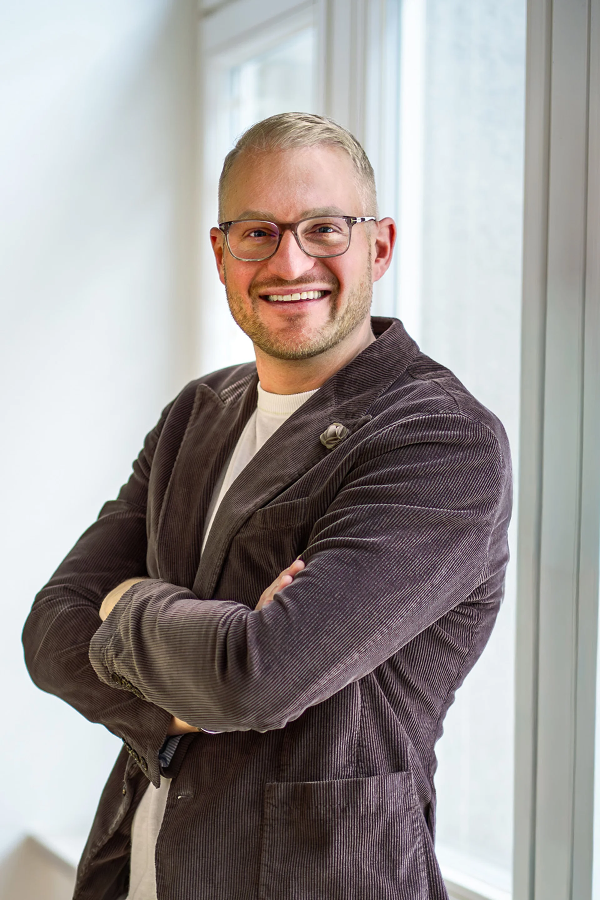 Person mit Brille und Jackett steht lächelnd vor einem Fenster.